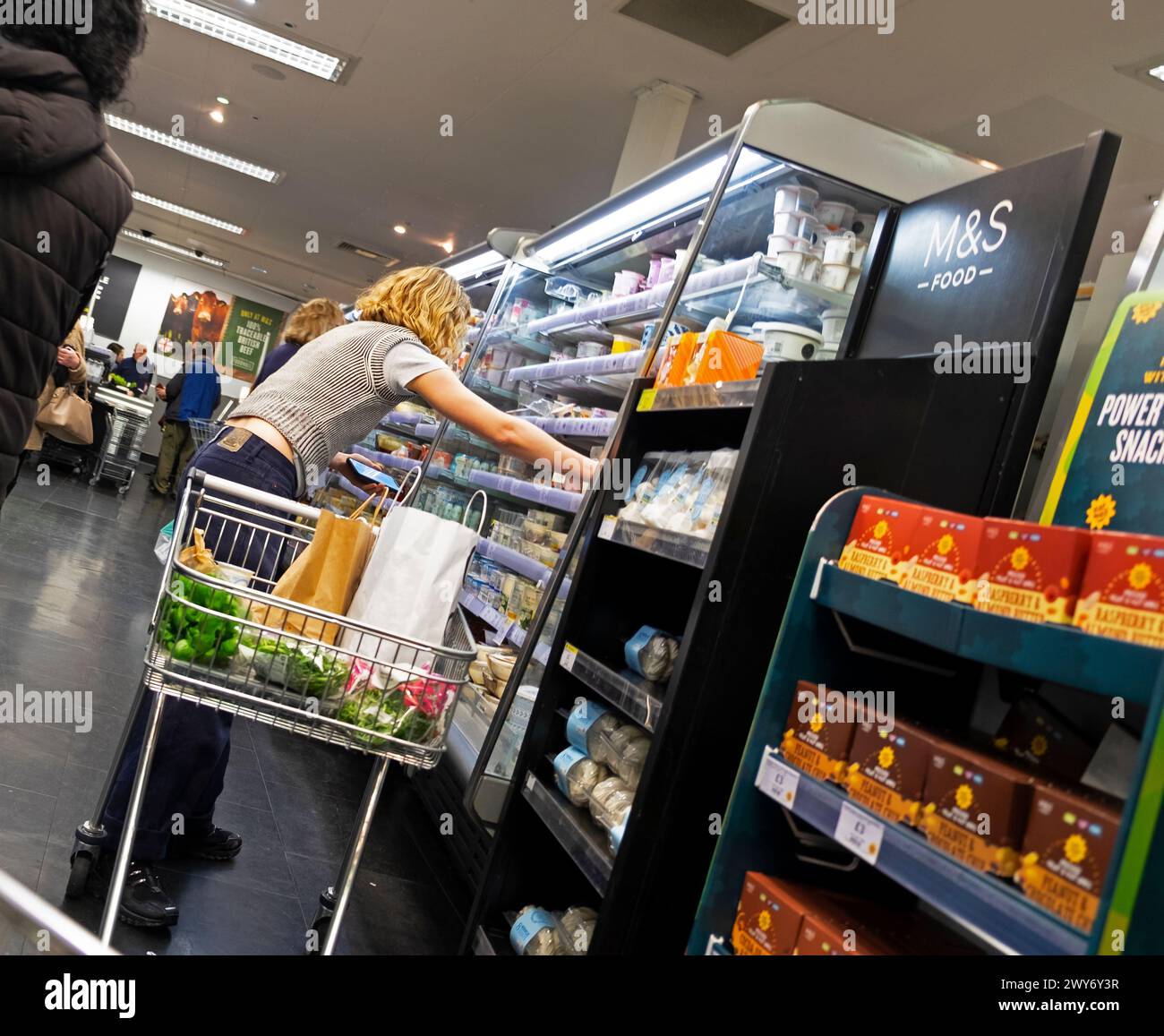 Einkaufswagen für junge Frau, Rückansicht, die nach einem Kühlregal für Joghurt in M&S Marks & Spencer Ladenhalle Innenraum 2024 UK KATHY DEWITT greift Stockfoto