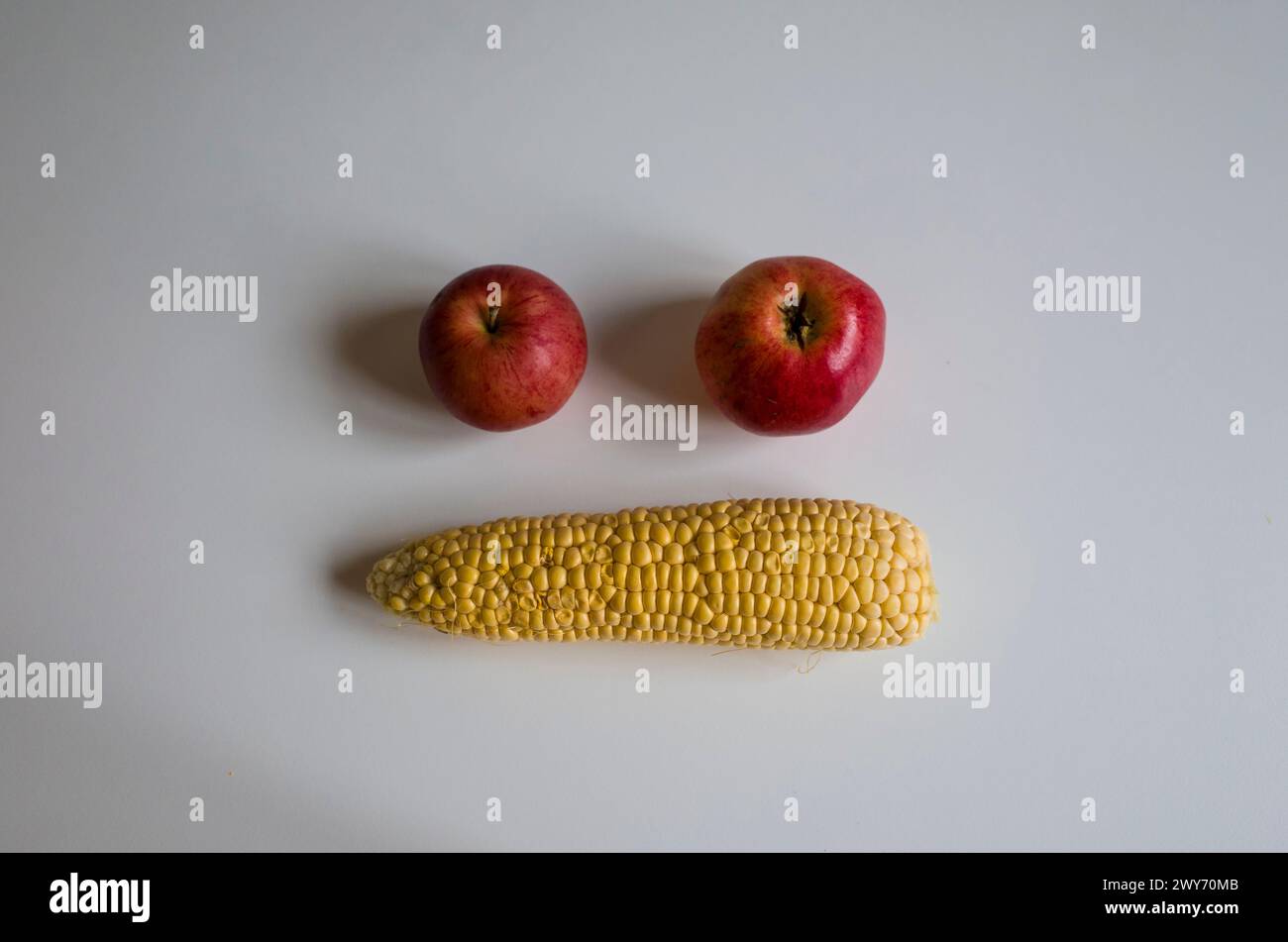 Gelb geschälte Maiskolben und zwei rote Äpfel bildeten Gesichtsemoji, zwei Augen und Mund auf weißem Tisch. Hausgemachtes echtes Bio-Gemüse nicht perfekt Stockfoto