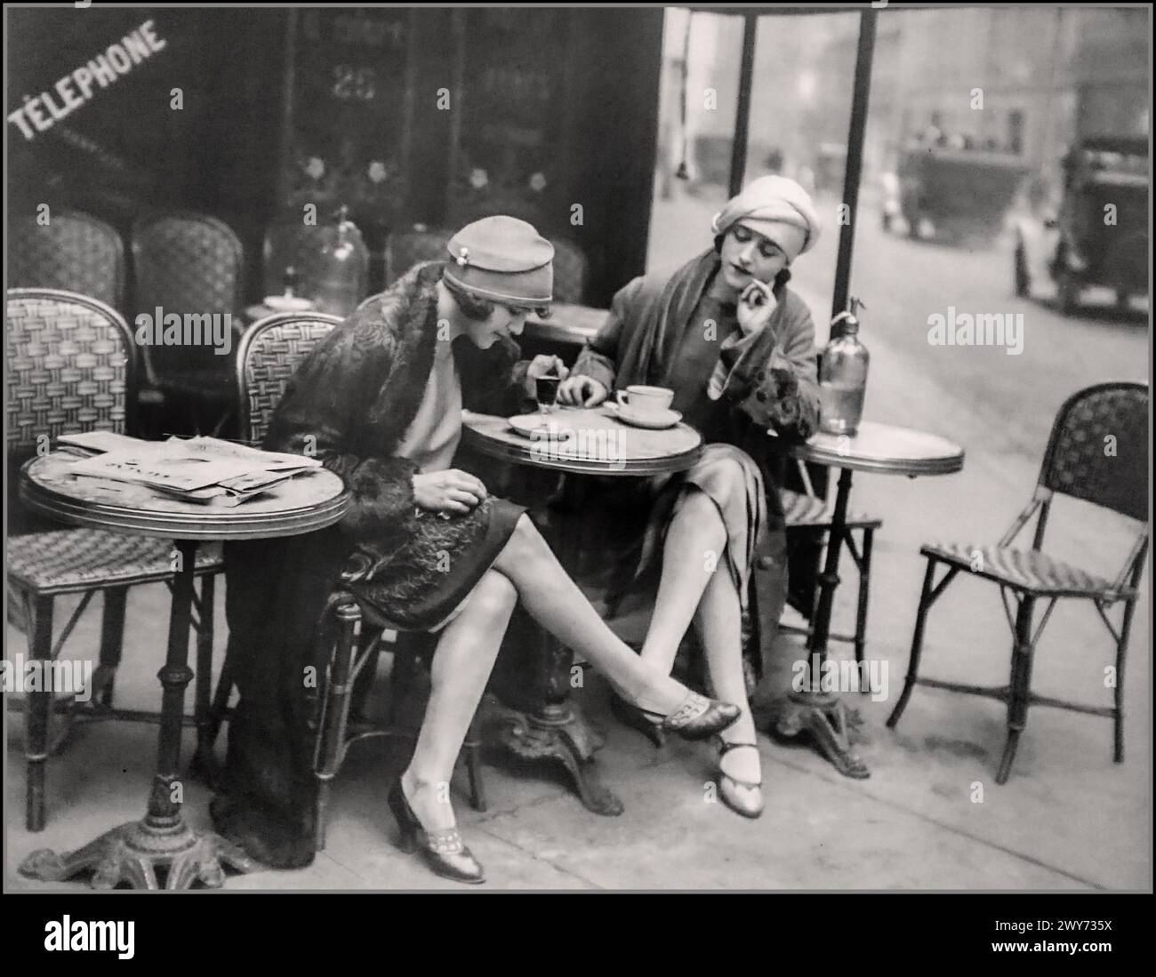 Vintage Paris Cafe 1920er Jahre, B&W zwei stilvolle modische Damen genießen Kaffee/Getränke in einem Boulevard Cafe Paris France 1920er Jahre Mode Architektur und Retro Lifestyle. Der Flapper Stil war der Stil, an den die meisten Leute denken, wenn sie an Mode der 1920er Jahre denken. Flapper-Frauen hackten ihr Haar zu einem kurzen, fast männlichen Stil ab. Sie trugen kurze, locker sitzende Kleider, kleinere Hüte wie Cloches, einen einfachen Hut, der wie die Glocke eines Blumentopfes geformt war. Stockfoto