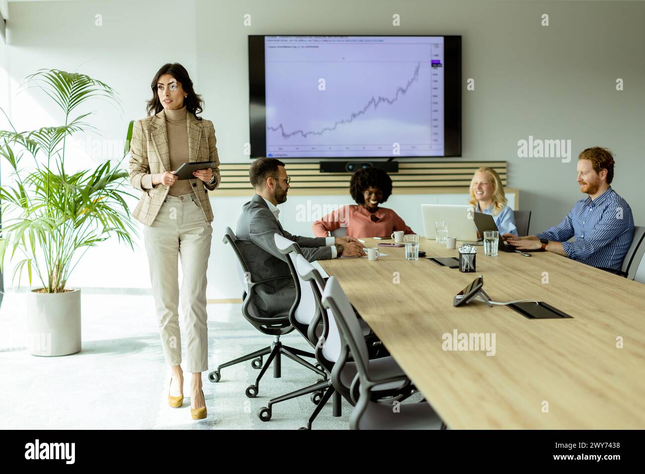 Entschlossene Führungskraft steht mit einem Tablet im Vordergrund und spricht ihr aufmerksames Team während eines Meetings an. Stockfoto