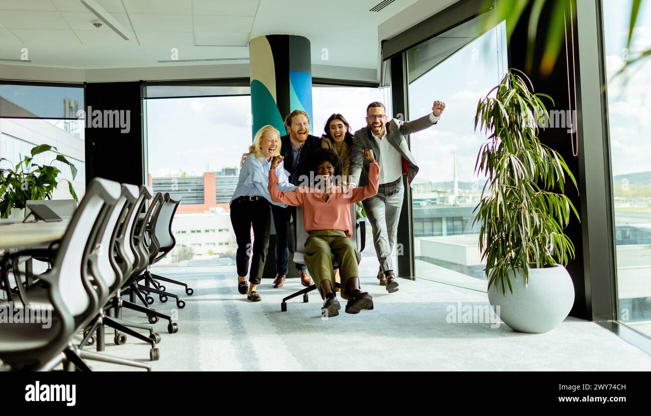 Ein Team von Kollegen teilt sich einen Moment des Spaßes und fährt mit einem Bürostuhl inmitten eines gut beleuchteten Arbeitsplatzes. Stockfoto