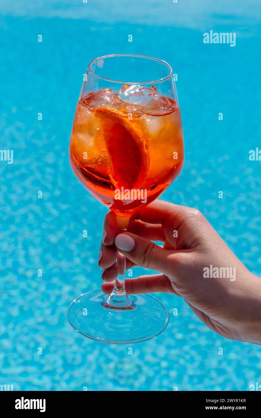 Aperol Spritz alkoholischer Cocktail im Glasbecher in der Hand der Frau mit weißem nagellack auf blauem Wasserpool Hintergrund Stockfoto
