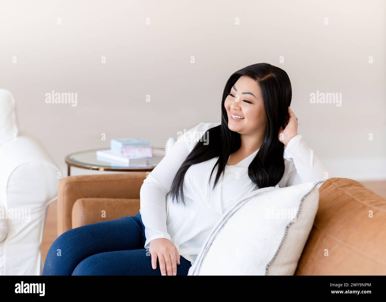 Asiatische Frau, die auf brauner Ledercouch sitzt und in der Ferne wegschaut Stockfoto