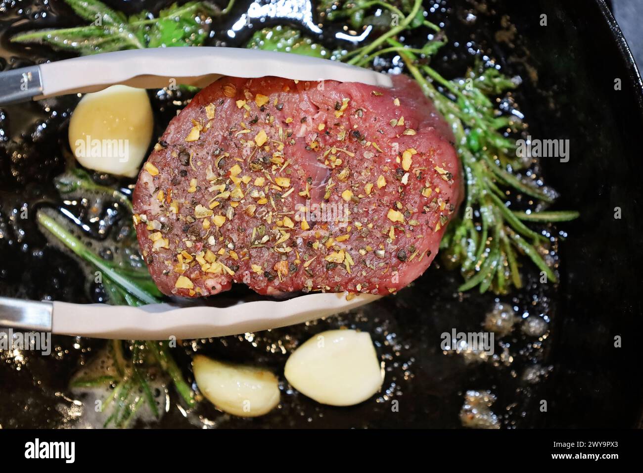 Zangen um ein Steak mit Kräutern, die in einer gusseisernen Pfanne anbraten Stockfoto
