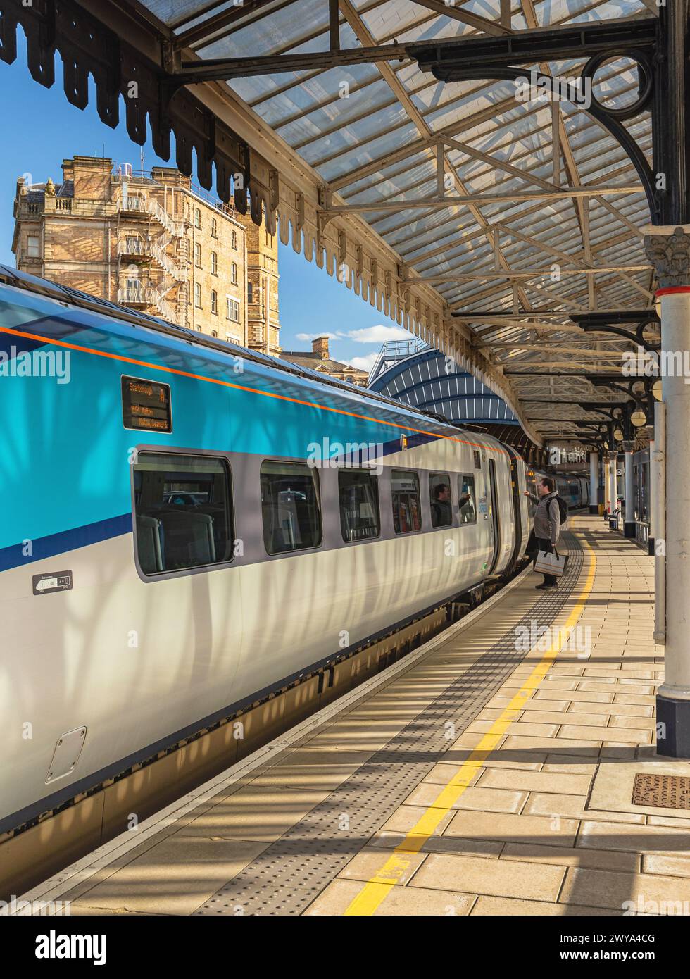 Ein Zug steht an einem Bahnsteig unter einem historischen Vordach aus dem 19. Jahrhundert. Architektonische Säulen stützen das Vordach und ein Hotelgebäude befindet sich im Hintergrund. Stockfoto