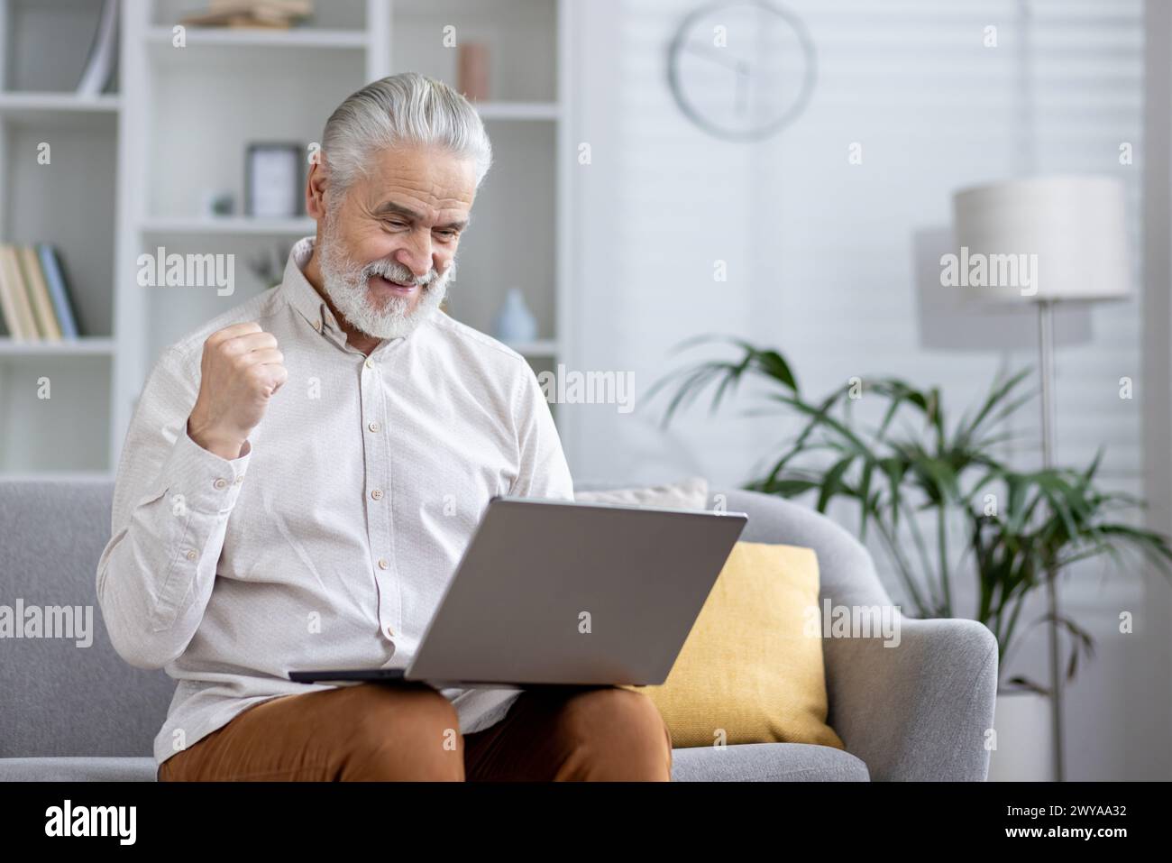 Ein älterer Herr, der einen Laptop benutzt und mit einer gewinnenden Geste in einem komfortablen Wohnzimmer begeistert ist. Stockfoto