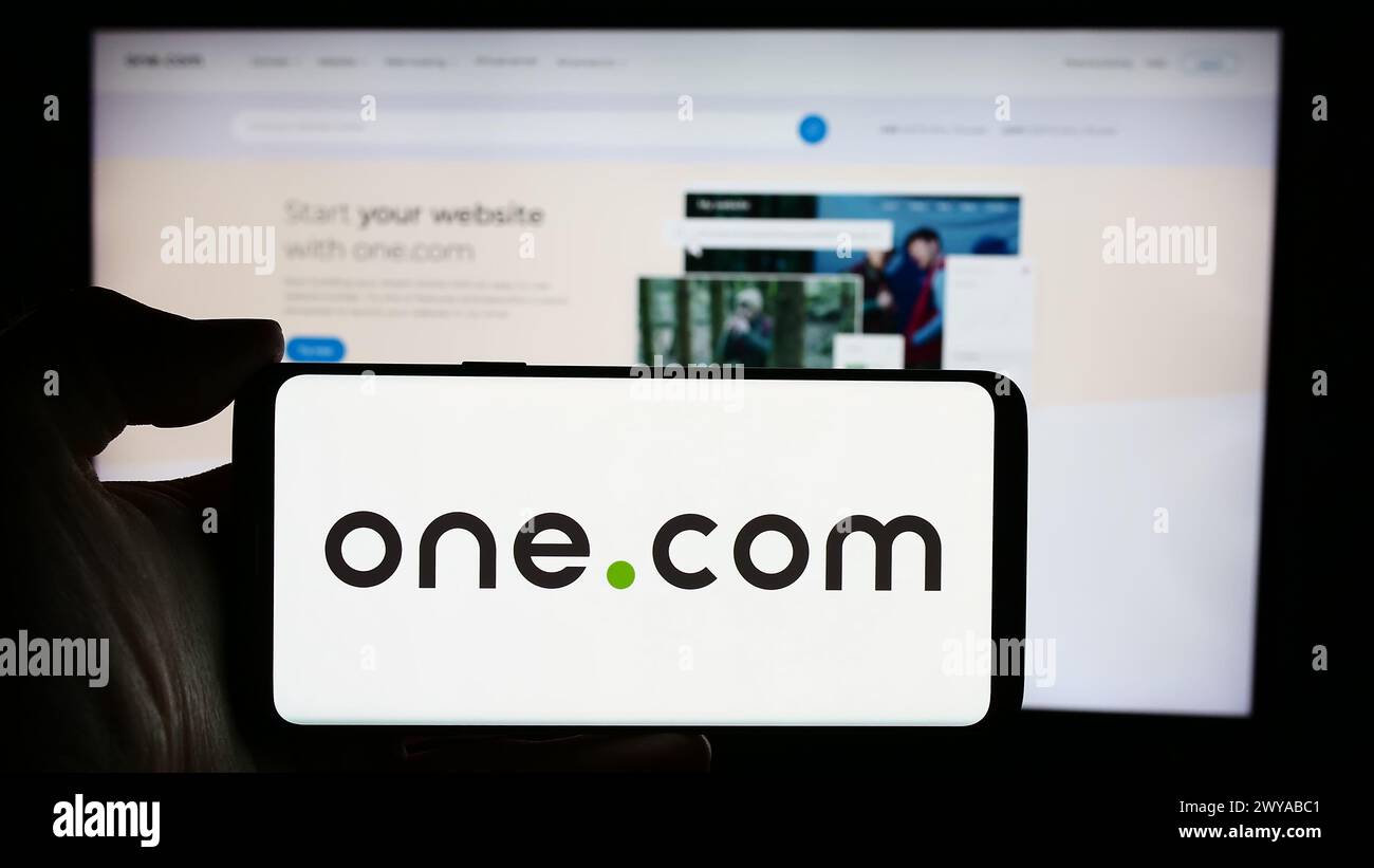 Person, die ein Mobiltelefon mit dem Logo des Webhosting-Unternehmens One.com Group vor der Business-Website hält. Schwerpunkt auf dem Display des Telefons. Stockfoto