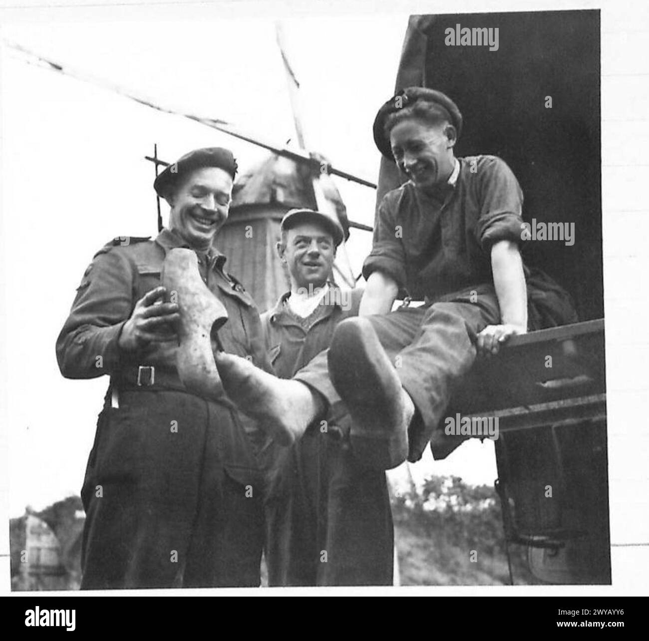 Britische Soldaten sind im Bild abgebildet, die während des Krieges in der Nähe von Windmühlen in den Niederlanden traditionelle niederländische Holzclogs anprobieren, wie von der britischen Armee, 21st Army Group dokumentiert. Stockfoto
