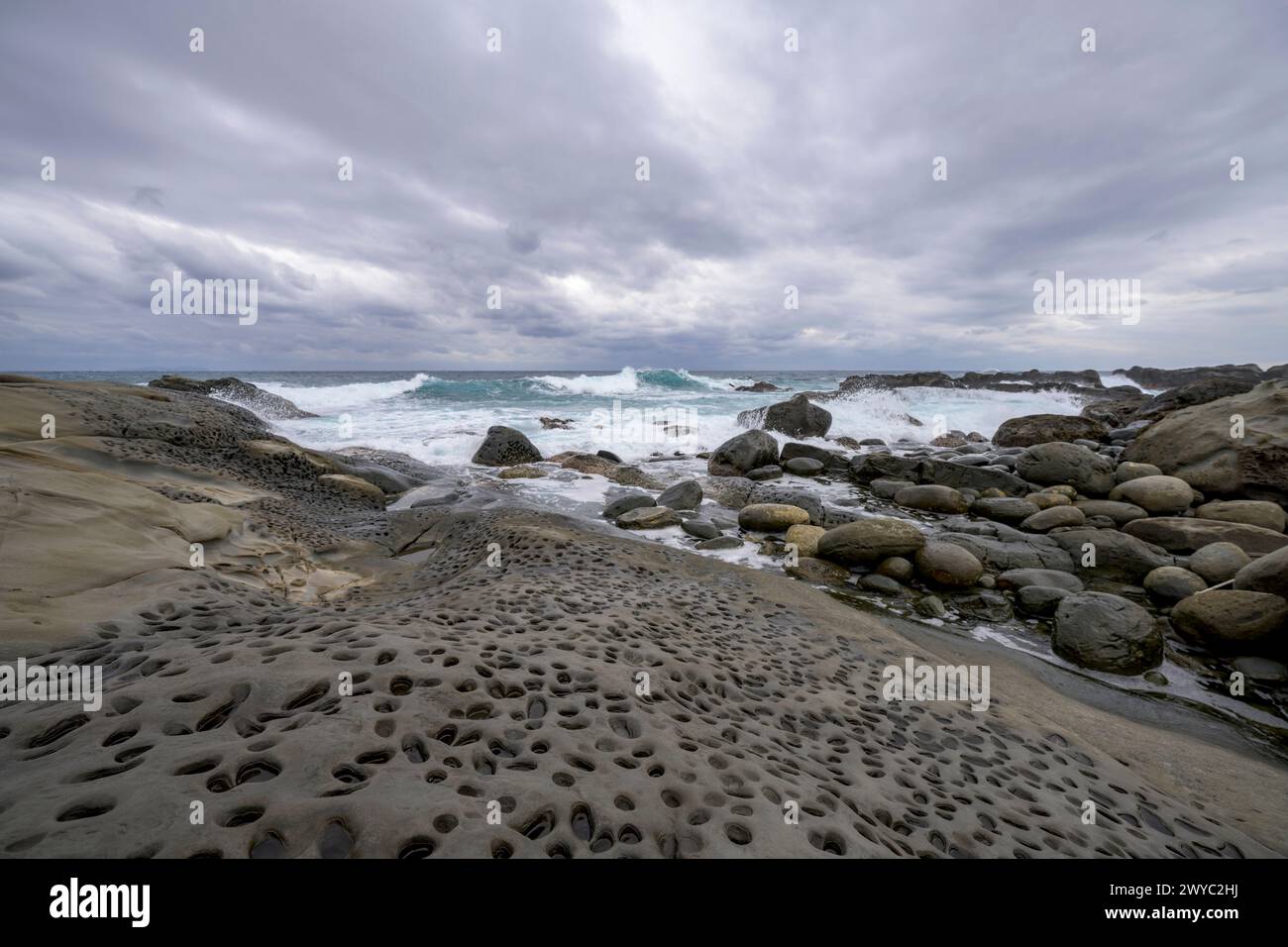 Ein bewölkter Tag an einem felsigen Ufer mit einzigartigen Erosionslochmustern mit rauen Wellen im Hintergrund Stockfoto