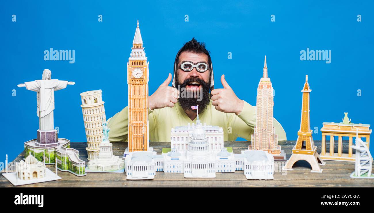 Glücklicher bärtiger Mann in Pilotenhelm und Brille mit Daumen nach oben in der Nähe des Tisches mit Miniaturmodellen. Miniaturkopie der Weltarchitektur Stockfoto