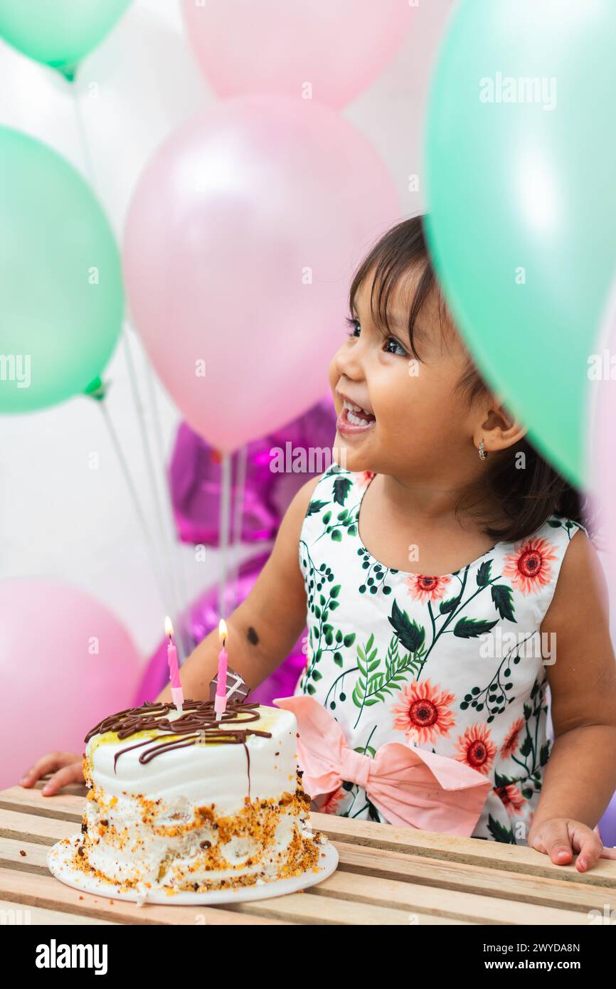 Nahaufnahme eines kleinen lateinischen Brünetten-Mädchens, das vor großem Glück vor ihrer Geburtstagskuche lächelt, umgeben von Ballons Stockfoto