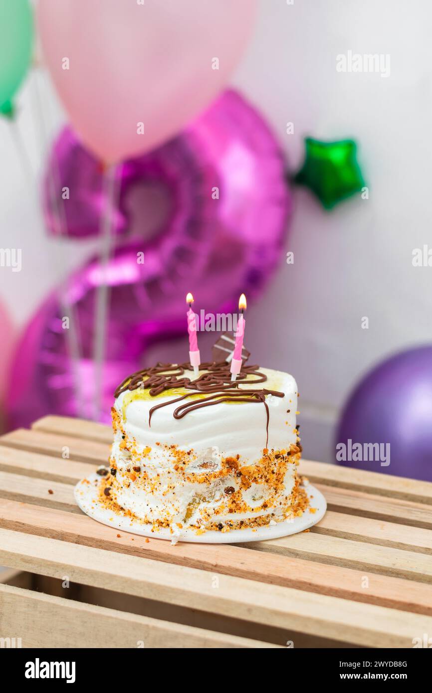 Kleiner Geburtstagskuchen mit zwei zündeten Kerzen darauf, auf einem Holztisch mit Ballons im Hintergrund Stockfoto