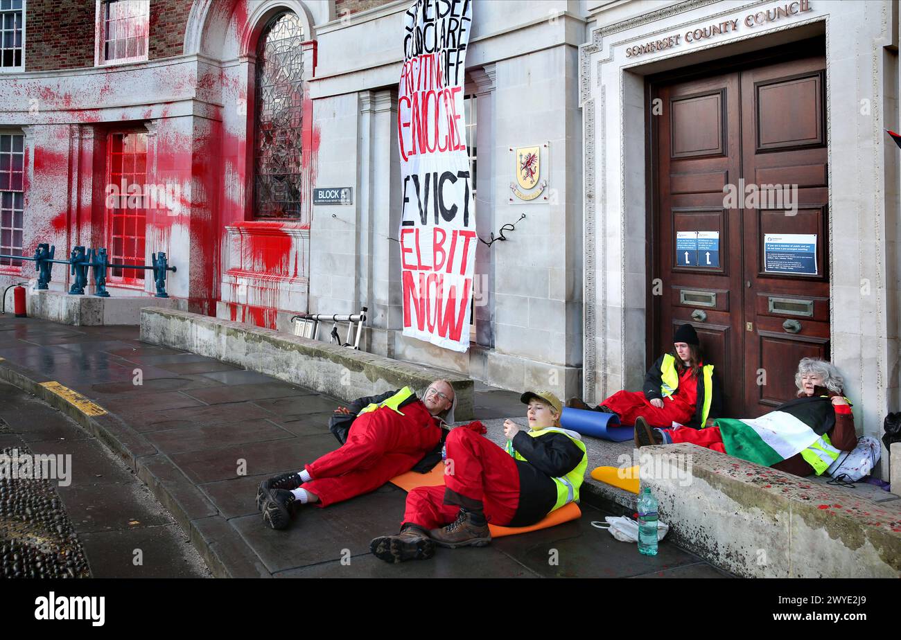 Palästinensische aktionäre sitzen in Zweiergruppen bei einer Demonstration vor dem Rathaus des Bezirks Somersetís. Unterstützer der Palästinensischen Aktion blockieren den Eingang zur County Hall mit Sperren und sprühen rote Farbe auf das Äußere des Gebäudes. Die Demonstranten fordern, dass der Stadtrat von Somerset den Mietvertrag für Büroflächen beendet, die sie an Elbit Systems in Aztec West, Bristol, vermieteten. Sie argumentieren, dass die von Elbit Systems in Großbritannien hergestellten Waffen von der israelischen Verteidigungsstreitkräfte gegen Palästinenser in Gaza und anderswo eingesetzt werden. Bei den israelischen Bombenanschlägen im Gazastreifen sind seit Oktober 2023 mehr als 30.000 Palästinenser ums Leben gekommen. Blass Stockfoto