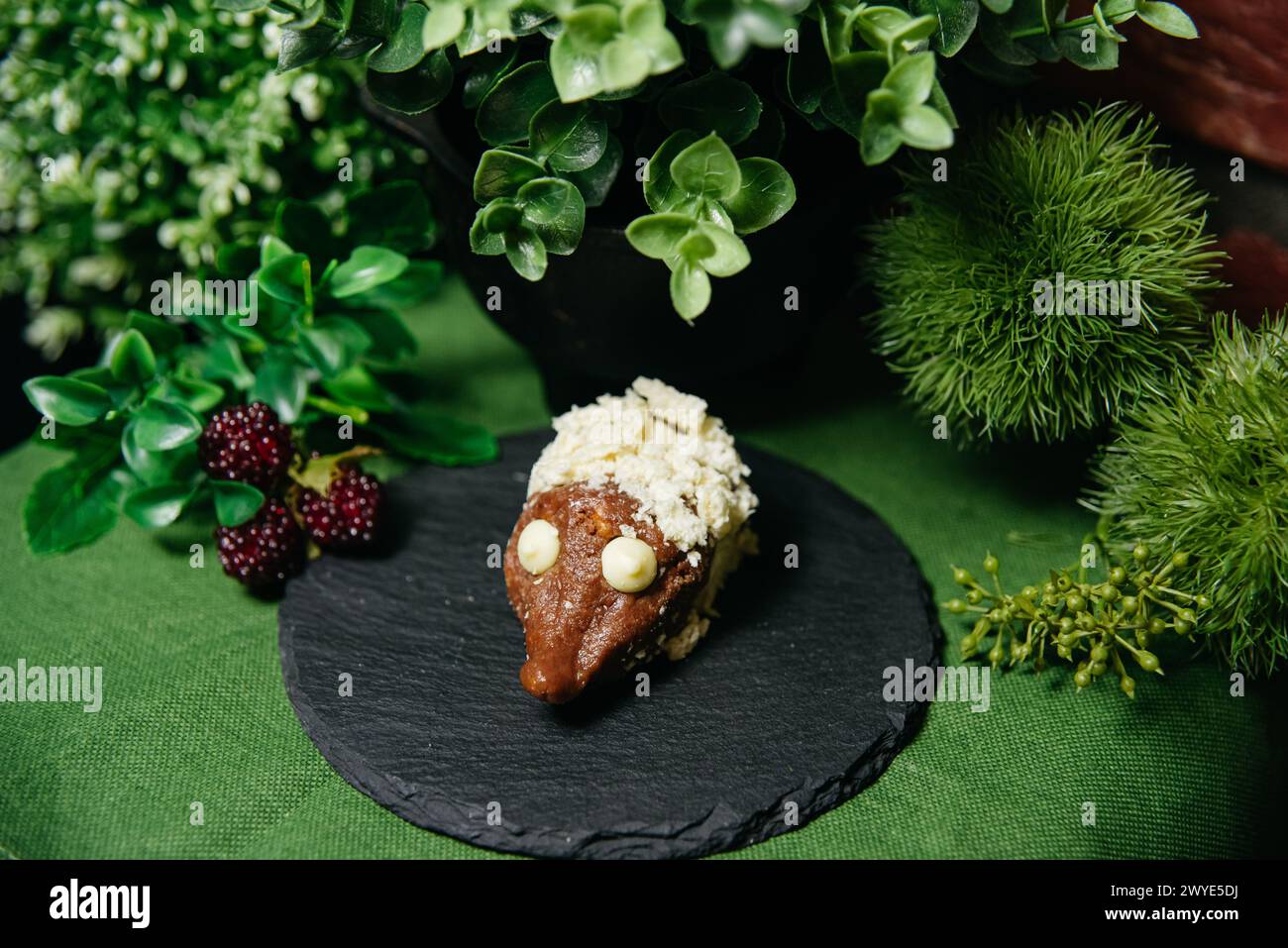 Ein dekadentes Gebäck in Form eines Igels, gekrönt mit Sahne und frischen Beeren, präsentiert auf einem Schieferbrett umgeben von üppigem Grün. Stockfoto
