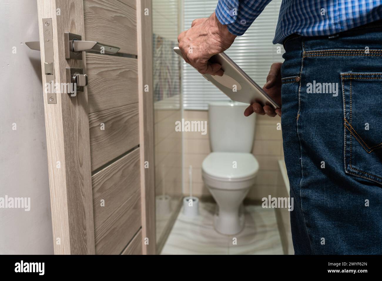 Ein älterer Mann in Jeans und Hemd steht über der Toilette und hält ein Klemmbrett. Stockfoto