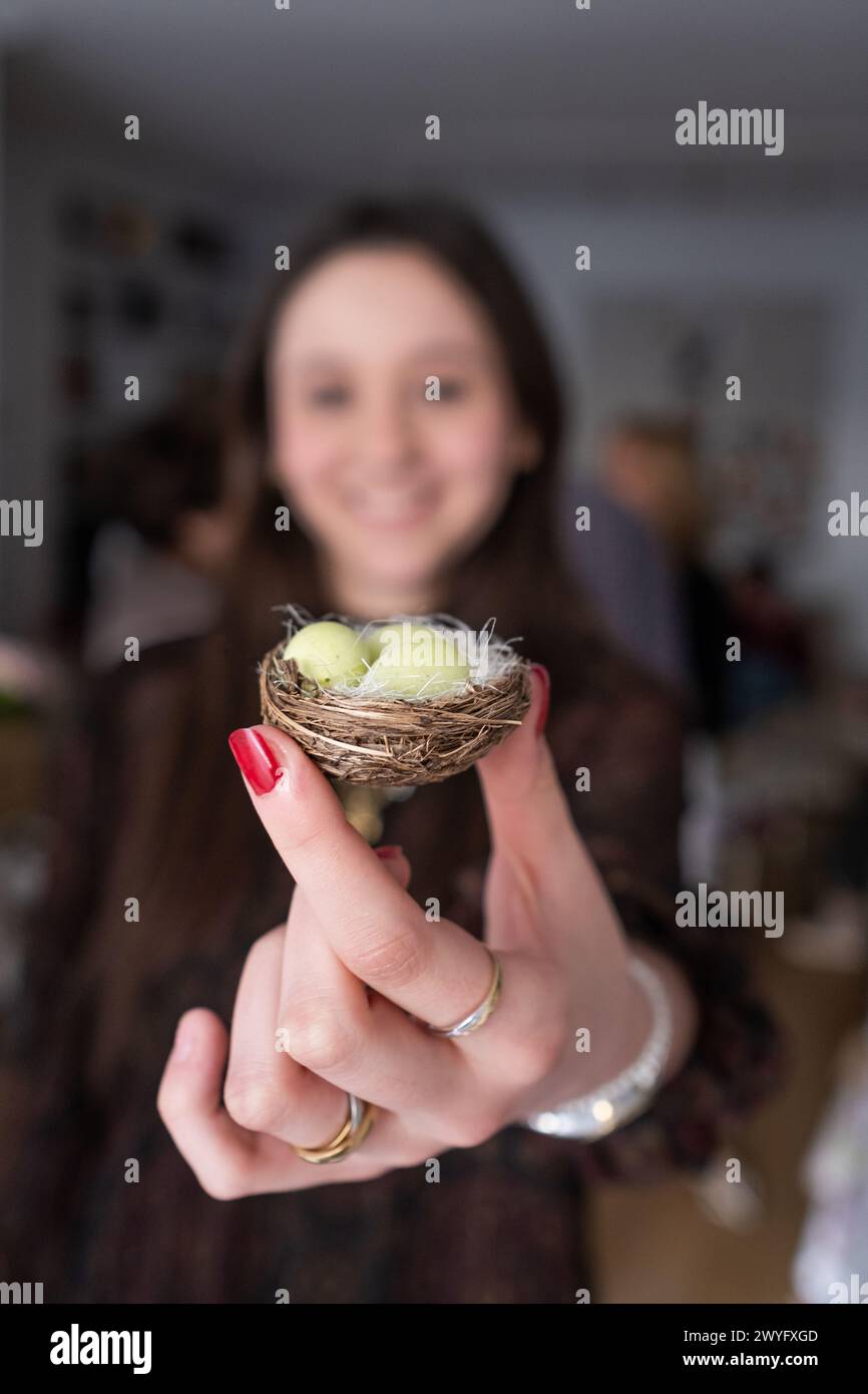 Eine lächelnde junge Frau hält vor der Kamera ein kleines Nest mit drei kleinen Schokoladen-Ostereiern, die grün bemalt sind Stockfoto