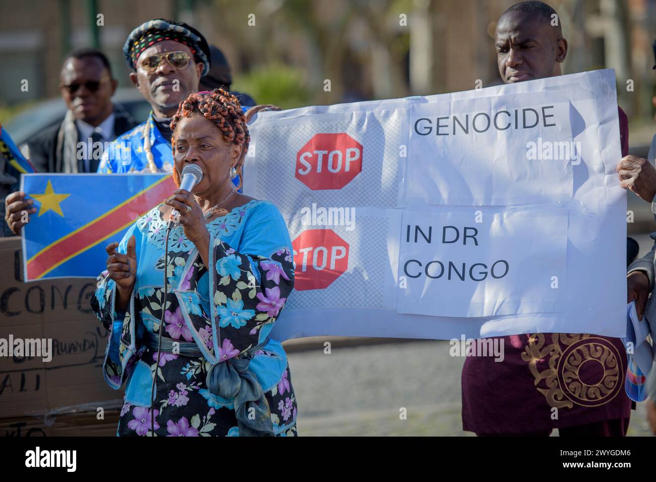 6. April 2024 in Rom, Italien: Demonstranten zeigen die Flagge des Kongo und ein Schild mit der Aufschrift "stoppt den Völkermord in der DR Kongo", während eine Frau in traditioneller afrikanischer Kleidung während der Demonstration in das Mikrofon spricht, um ihre Solidarität mit dem kongolesischen Volk auszudrücken. die Forderung nach einem "Stopp des anhaltenden Völkermords" und einem Ende der Ausbeutung des afrikanischen Kontinents, die von einigen Verbänden der afrikanischen Gemeinschaften Europas zusammen mit dem Kommunistischen Netzwerk und der Bewegung Hands Off from Africa in Rom organisiert werden. 30 Jahre bewaffneter Konflikt finanziert durch die westlichen W Stockfoto