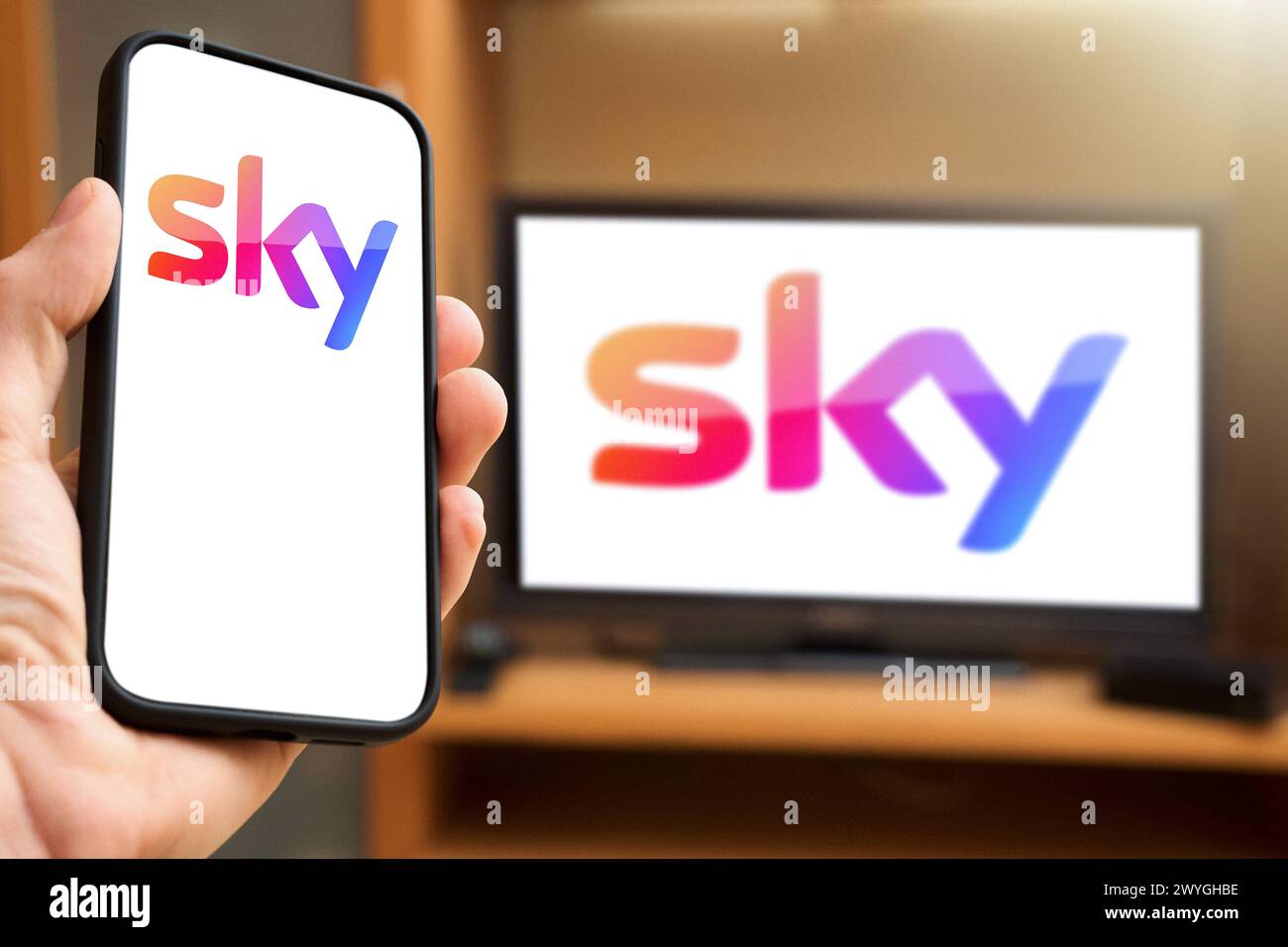 6. April 2024: Smartphone mit Sky Pay TV und Streaming Provider-Logo vor dem Fernsehbildschirm halten. FOTOMONTAGE *** Handheld Smartphone vor TV Bildschirm mit dem Pay-TV und Streaming Anbieter Logo von Sky. FOTOMONTAGE Stockfoto