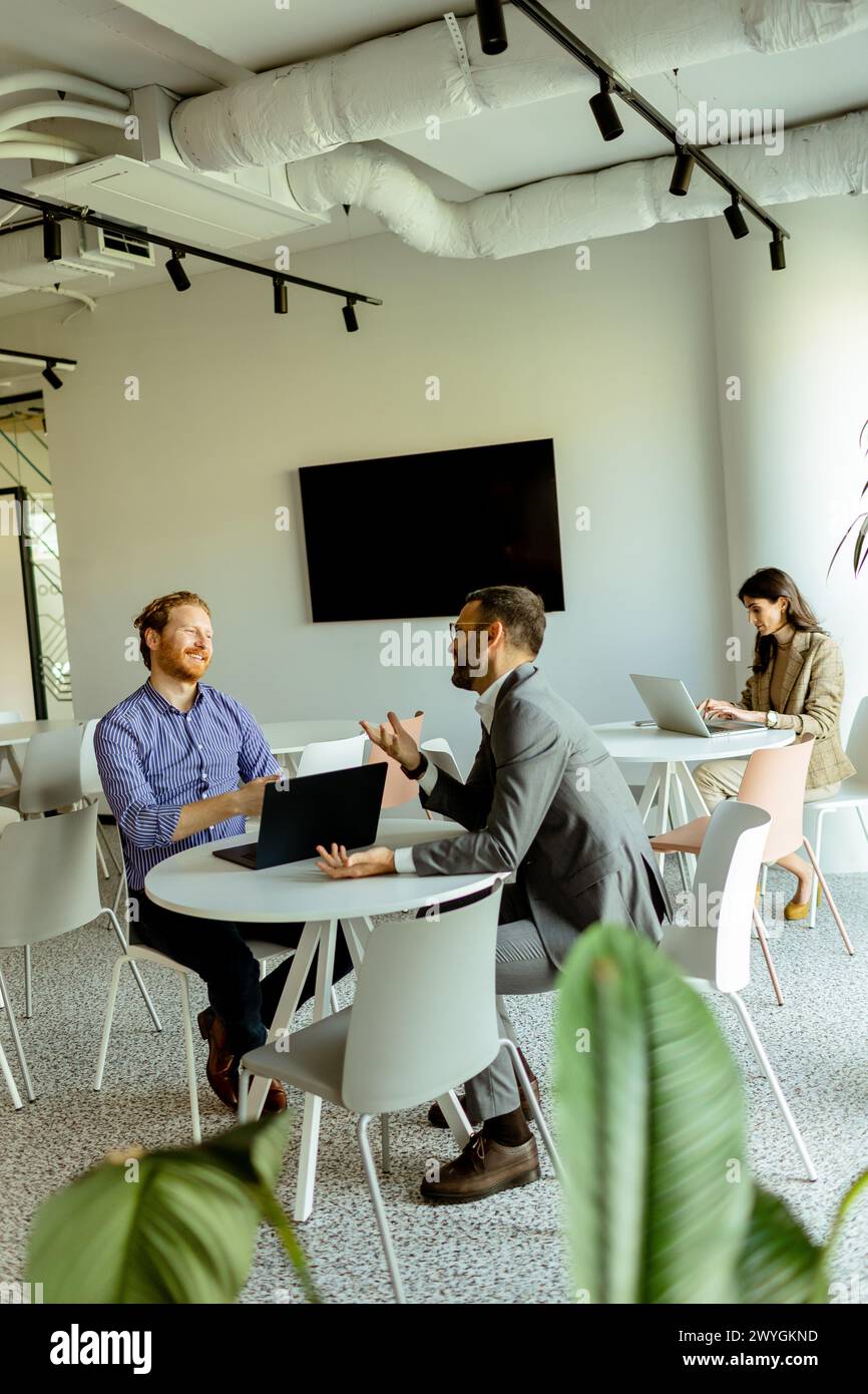 Zwei Männer führen ein lebhaftes Gespräch an einem Tisch, während eine Frau im Hintergrund unabhängig arbeitet Stockfoto