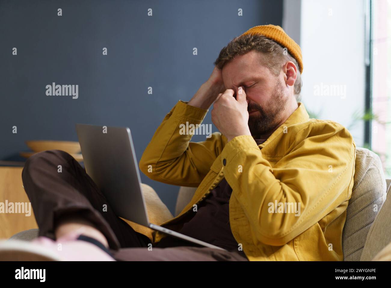 Ein Mann sitzt auf einem Stuhl mit einem Laptop auf dem Schoß. Er trägt eine gelbe Jacke und eine gelbe Mütze. Er schaut auf den Laptop-Bildschirm mit einem traurigen Stockfoto