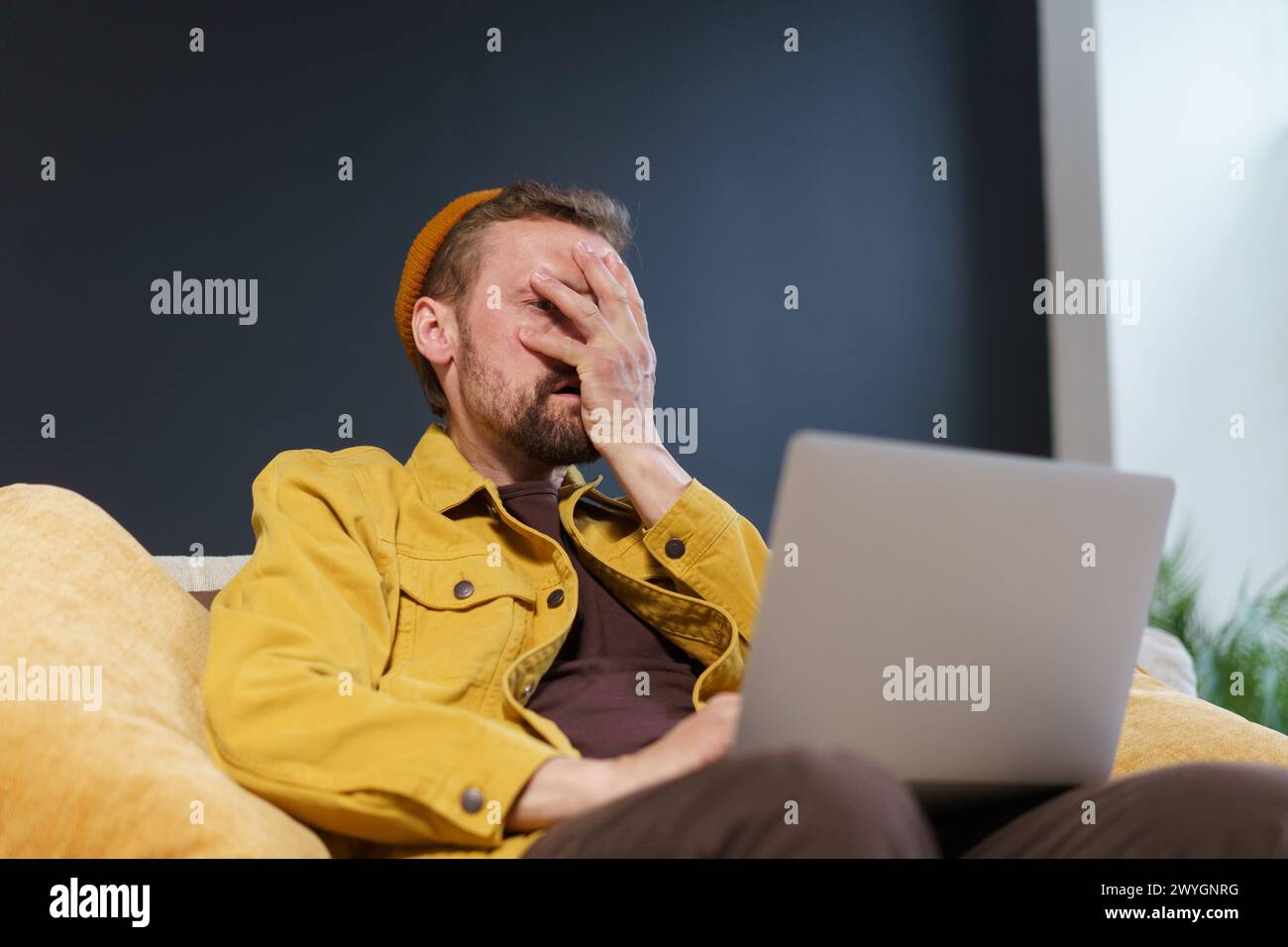 Ein Mann sitzt auf einer Couch mit einem Laptop vor sich. Er trägt eine gelbe Jacke und einen Hut. Er schaut sich den Laptop mit einem verwirrten Verfall an Stockfoto