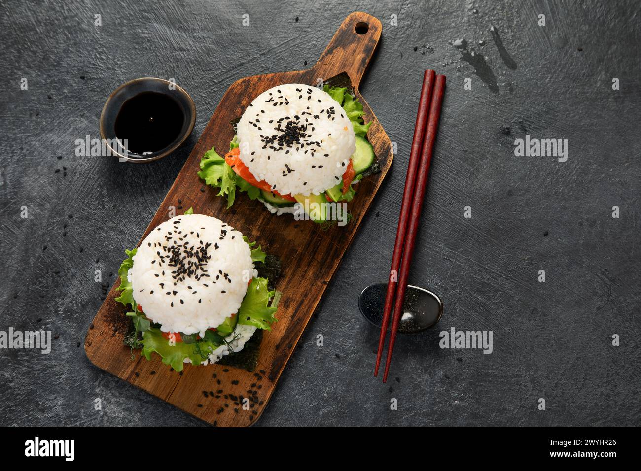 Asiatischer Sushi-Burger, Sojasauce, Essstäbchen auf dunklem Hintergrund. Trendiges Hybrid-Essen. Blick von oben Stockfoto