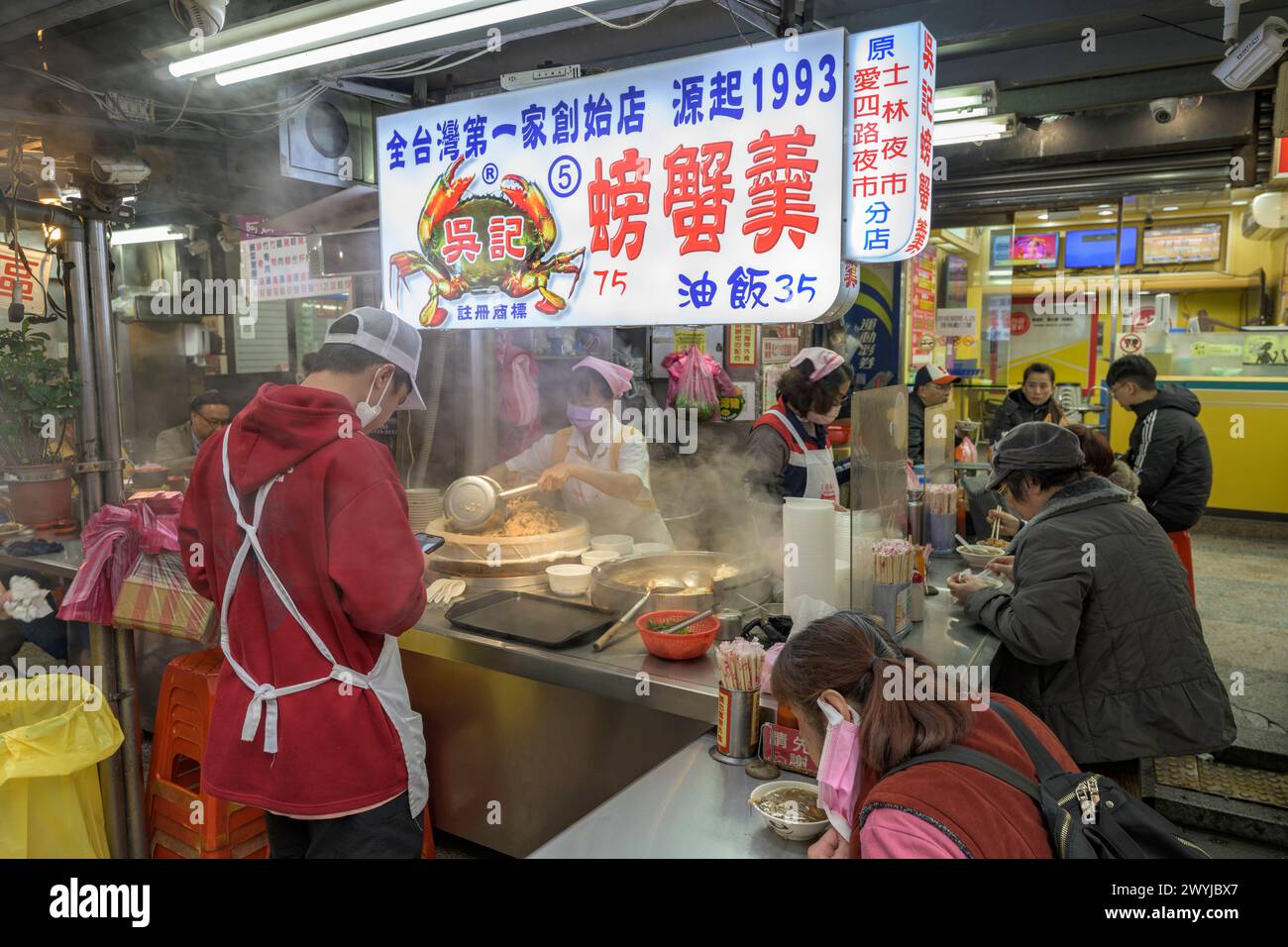 Nachtmarktszene mit Köchen, die kochen, und Gästen, die an einem belebten Imbissstand mit bunten Schildern sitzen Stockfoto