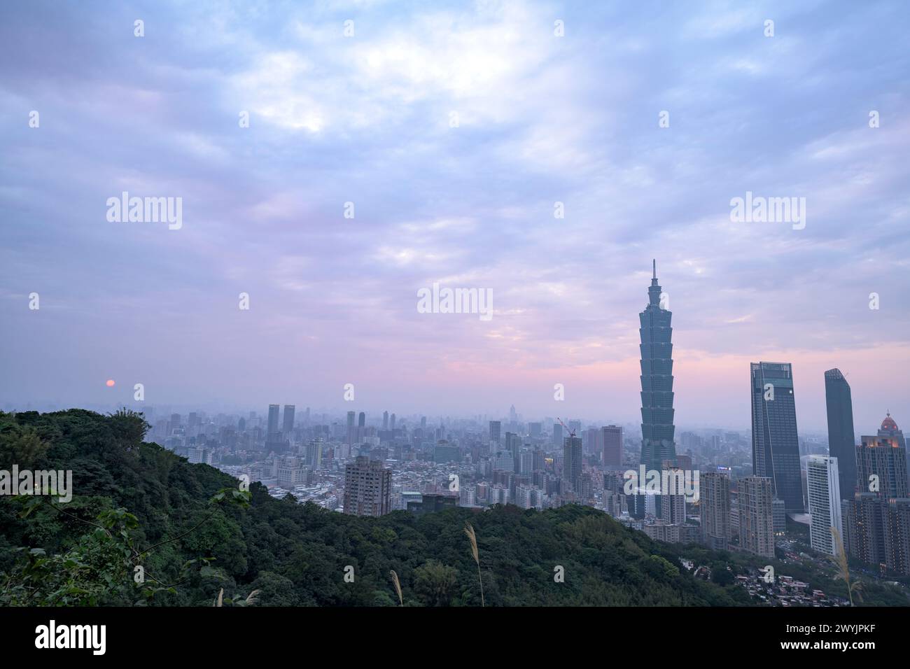 Ein erhöhter Blick auf Taipeh Stadt während der Dämmerung mit Wolkenkratzern vor einem subtilen Sonnenuntergang, aufgenommen von einem bergigen Gebiet mit Laub Stockfoto