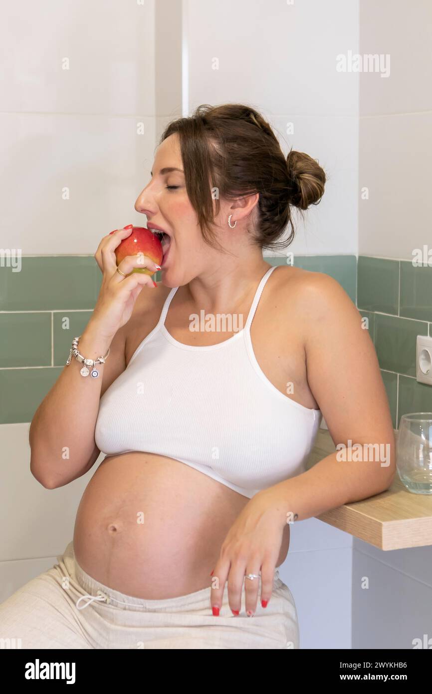 Schöne schwangere Frau in bequemen Kleidern, die einen roten Apfel in der Küche isst. Stockfoto