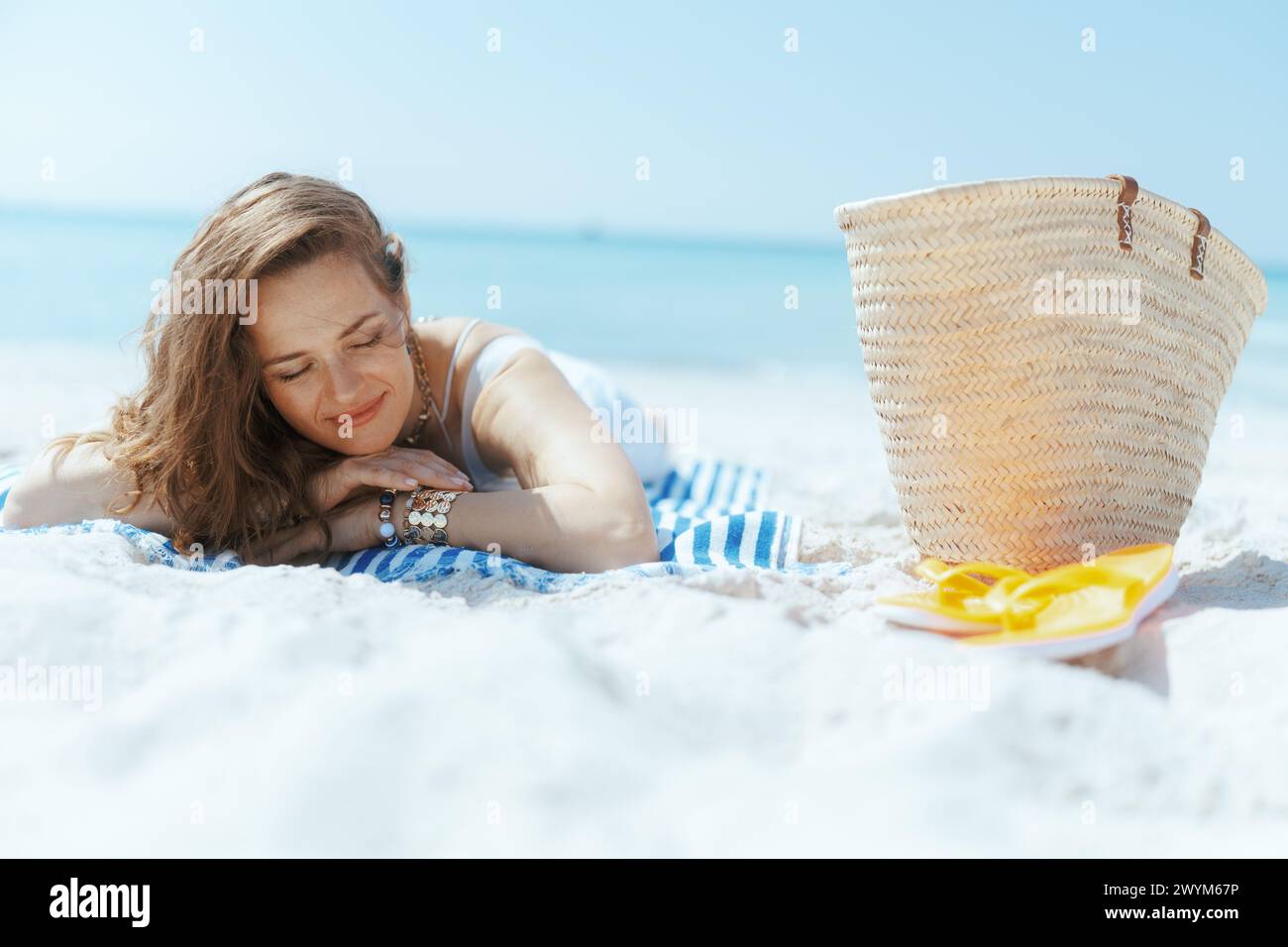 Entspannte elegante 40-jährige Frau am Meer mit Strohbeutel und gestreiftem Handtuch. Stockfoto