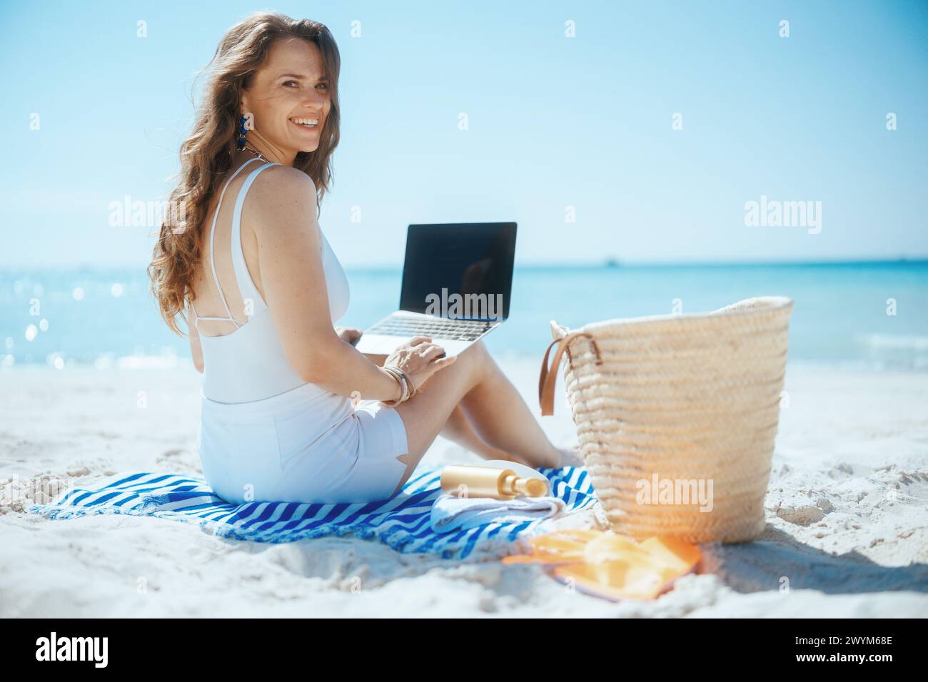 Glückliche, elegante Frau an der Meeresküste mit Strohtasche, leerem Laptop-Bildschirm und gestreiftem Handtuch. Stockfoto