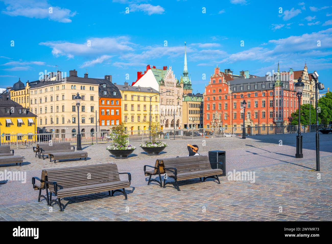 Bänke säumen einen kopfsteingepflasterten Platz in Stockholm, umgeben von historischen, farbenfrohen Gebäuden unter einem klaren blauen Himmel. Schweden Stockfoto