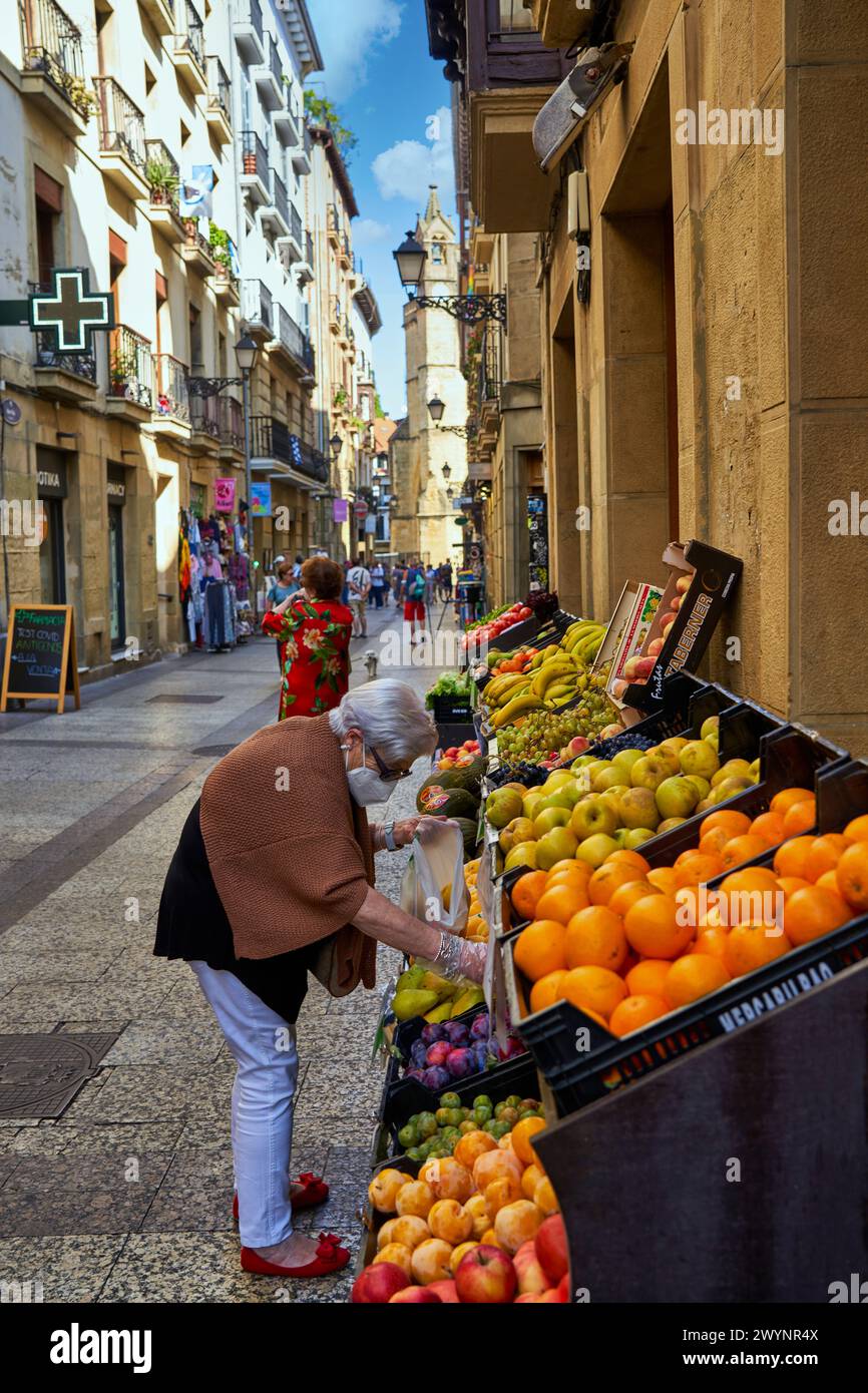 Mujer comprando en frutería de la parte Vieja, al fondo Iglesia de San Vicente, Donostia, San Sebastián, Gipuzkoa, País Vasco, España, Europa, Adentrarse en la parte Vieja es conocer el verdadero centro Social de Donostia, sus calles empedradas como la del 31 de Agosto, que conmemora el Incendio ese día de 1813, están llenas de bares para disfrutar de los mejores pintxos de la ciudad, y sus edificios emblemáticos como la Basílica de Santa María del Coro y la iglesia de San Vicente Son de gran atractivo e interés,. Stockfoto