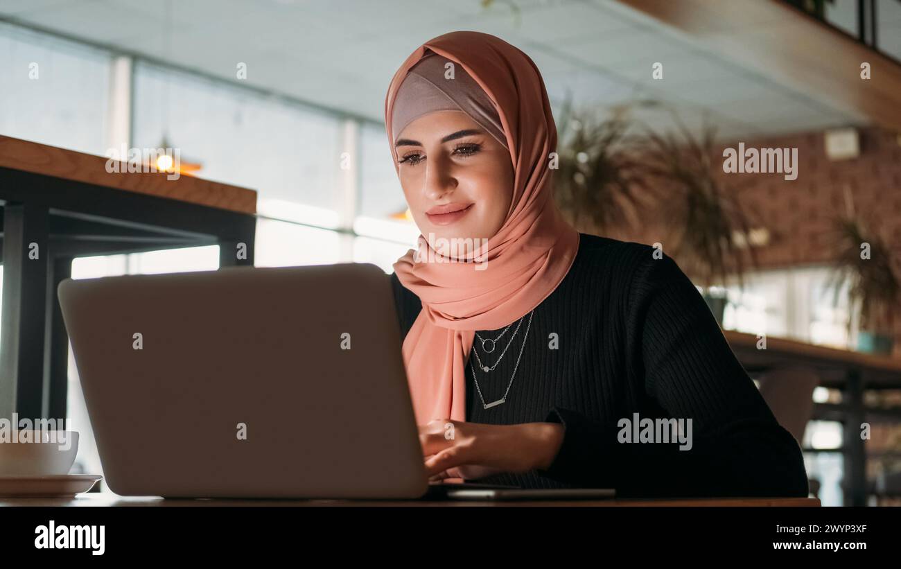 Fernarbeit. Freiberuflicher Mitarbeiter. Lächelnde, konzentrierte Geschäftsfrau im Hijab, die auf einem Laptop im Café im Internet surft. Stockfoto