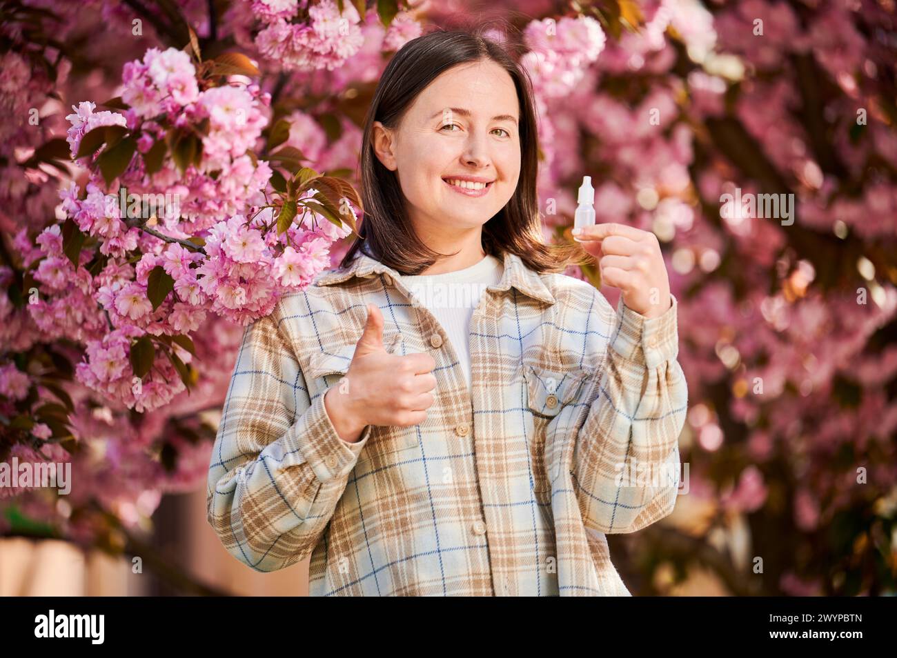 Frau, die medizinische Augentropfen verwendet, leidet unter saisonaler Allergie im Frühling im blühenden Garten. Porträt einer glücklichen Frau, die die Daumen hoch vor einem blühenden Baum zeigt. Frühjahrsallergiekonzept. Stockfoto