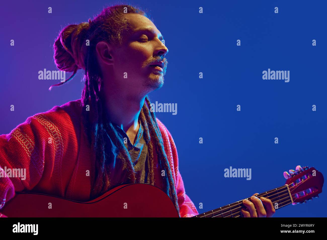 Fesselnder Anblick eines Mannes mit Dreadlocks, der seine Seele in Musik gießt, Gitarre vor blauem Hintergrund im Neonlicht spielt Stockfoto