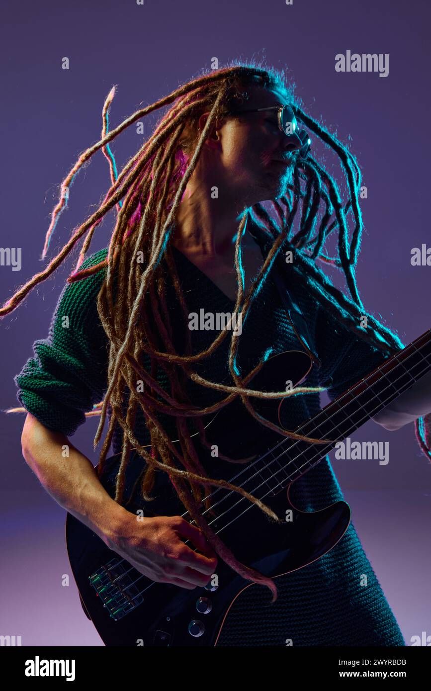 Leidenschaftlicher Darsteller mit Dreadlocks, die sein Herz durch seine Gitarre ausgießen und vor dunklem lila Hintergrund im Neonlicht spielen Stockfoto