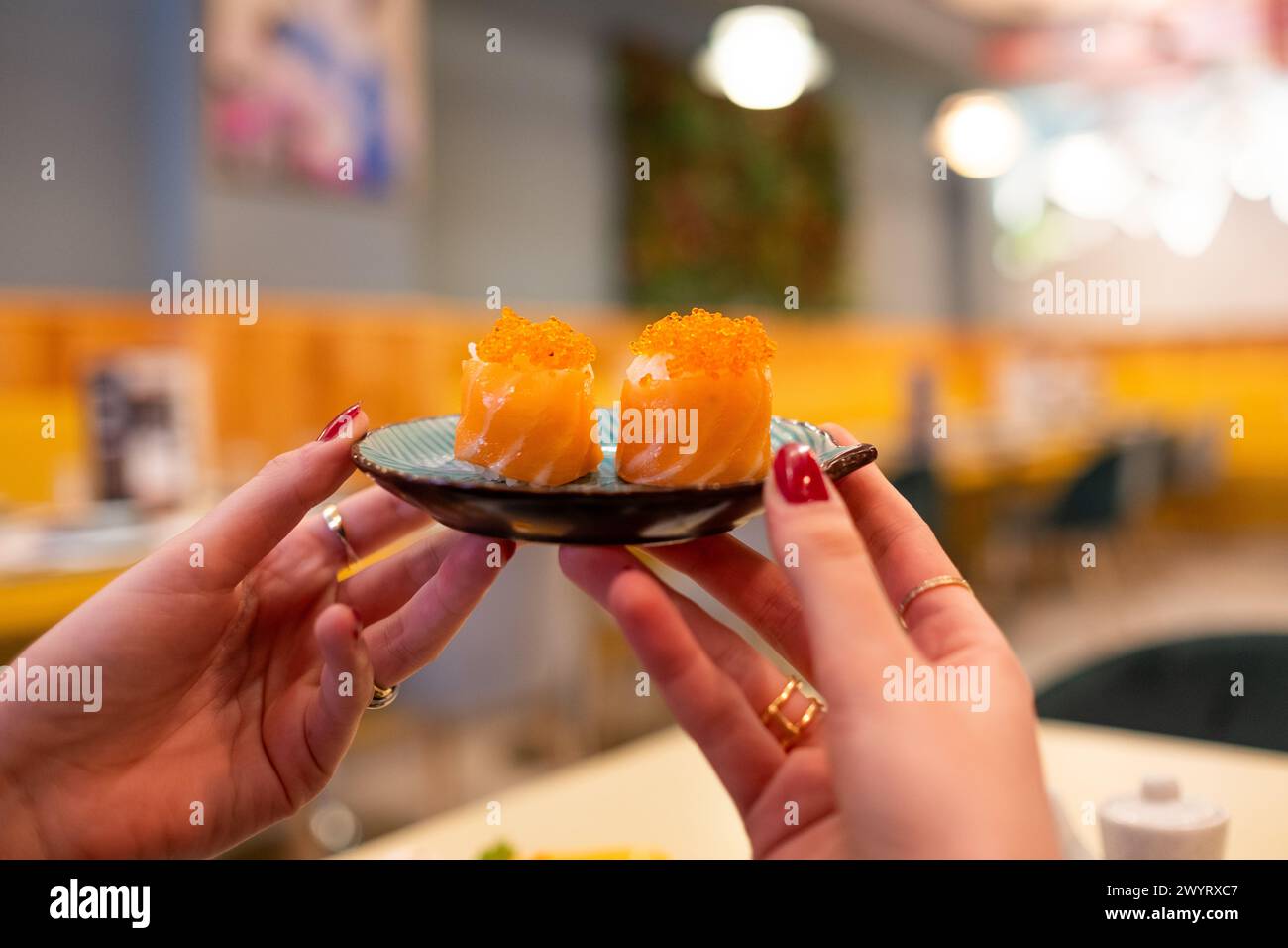 Eine Hand hält einen kleinen Teller mit zwei Lachs-Maki-Rollen vor einem warmen und farbenfrohen japanischen Restaurant vor der Kulisse. Das Bokeh und die Hintergrundunschärfe sind sehr Stockfoto