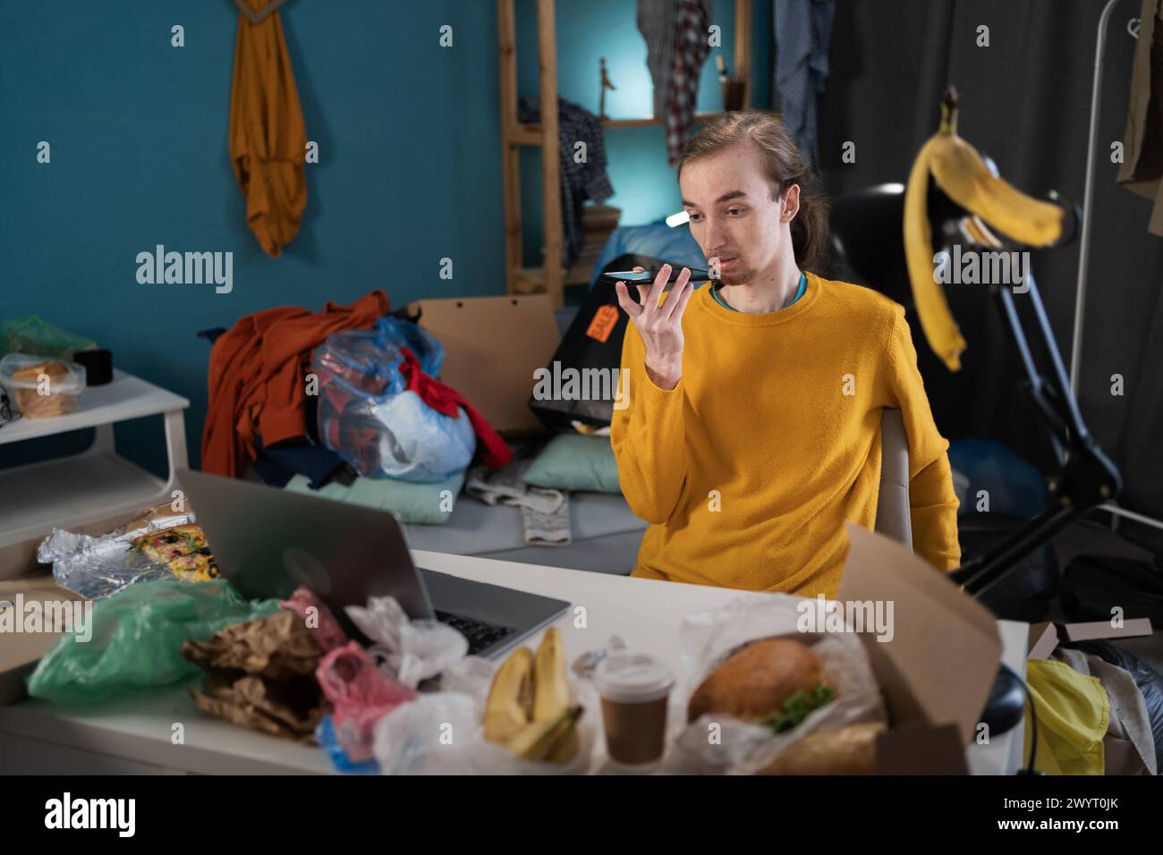 Junger Mann, der in einem unordentlichen Zimmer studiert, diktiert Sprachnachrichten auf dem Smartphone Stockfoto