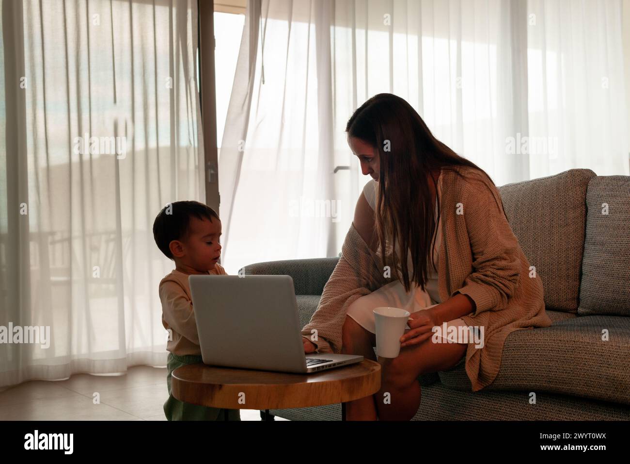 Mama spricht mit Baby, während sie zu Hause am Laptop arbeitet, Kaffeetasse in der Hand. Stockfoto