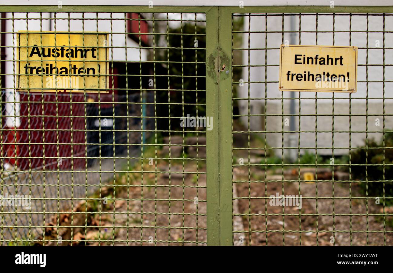 Gelbe Warnschilder auf Deutsch an einem grünen Zauntor mit der Aufschrift „Keep Entry and Exit Clear!“ („Ausfahrt freihalten“, „Einfahrt freihalten“) Stockfoto