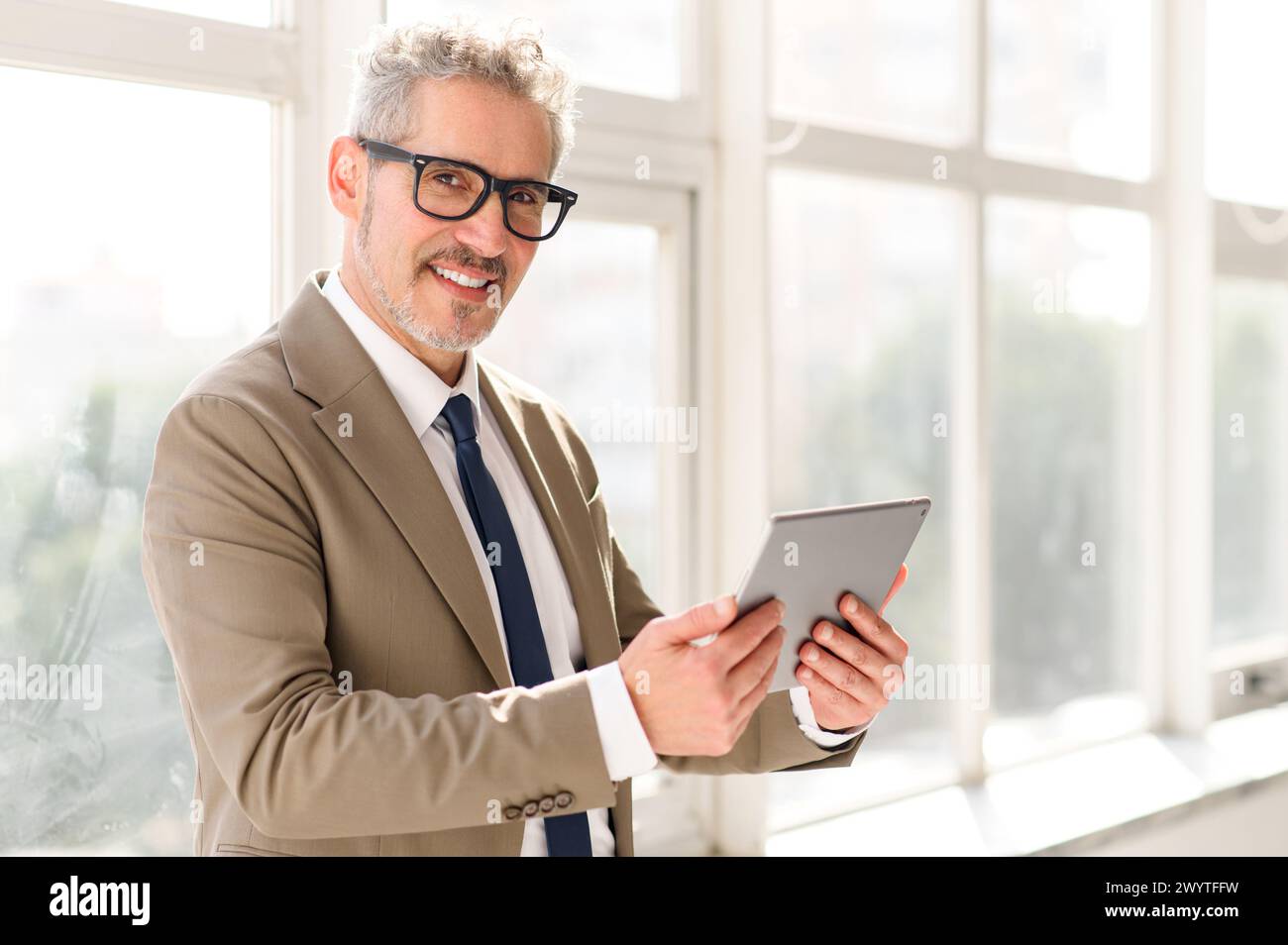 Die leitende Führungskraft, die in einem Raum mit Tageslicht und großem Fensterblick steht, navigiert mühelos durch ein Tablet und zeigt die nahtlose Integration von Technologie in das moderne Geschäftsleben. Stockfoto