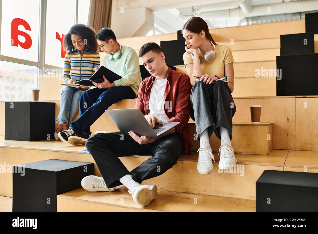Eine multikulturelle Gruppe von Schülern sitzt auf Stufen und arbeitet in einer Bildungsumgebung zusammen an einem Laptop. Stockfoto