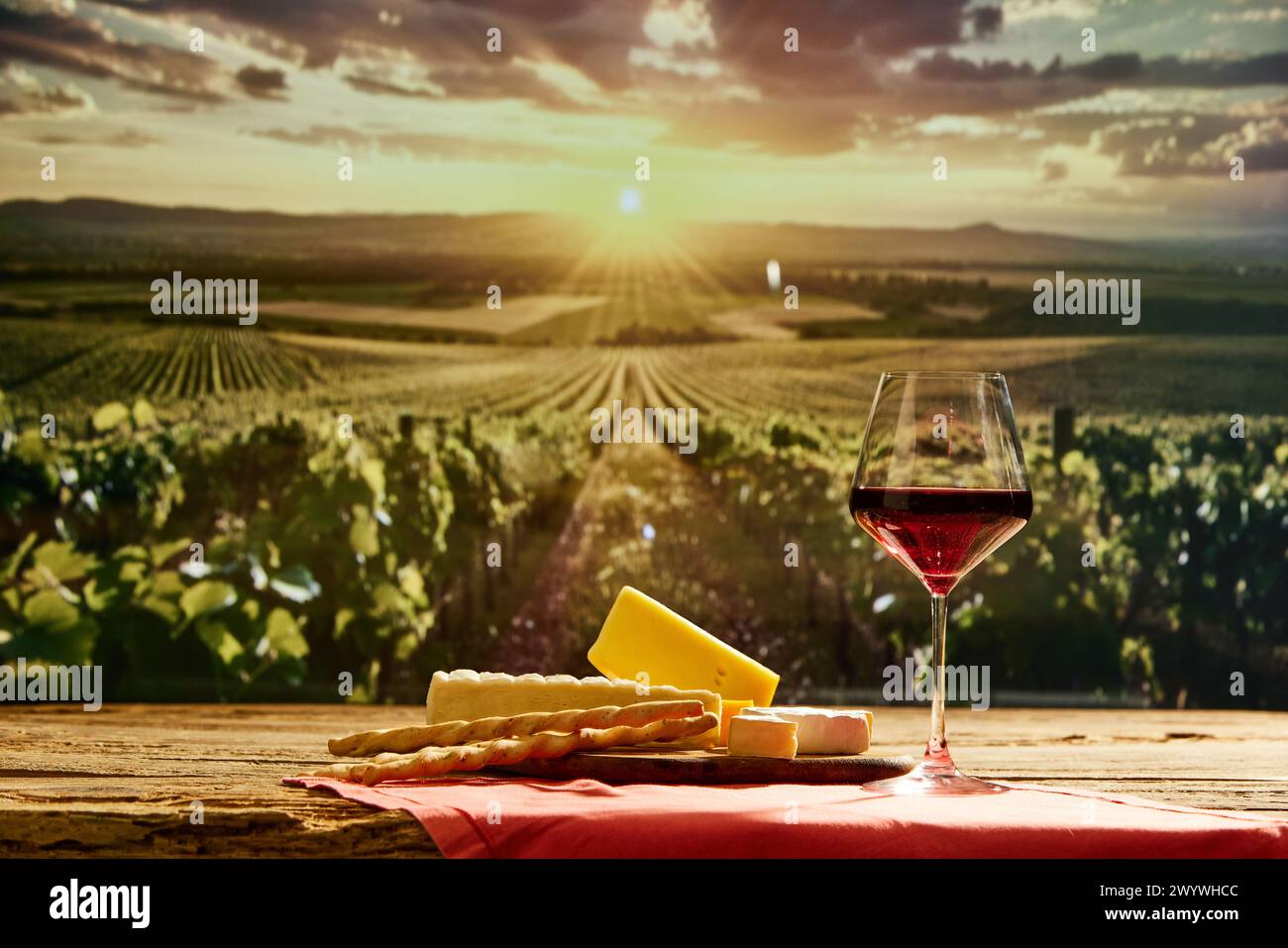 Glas Rotwein auf Holztisch mit Käse und Brotstangen mit Weinberg während Sonnenuntergang im Hintergrund Stockfoto