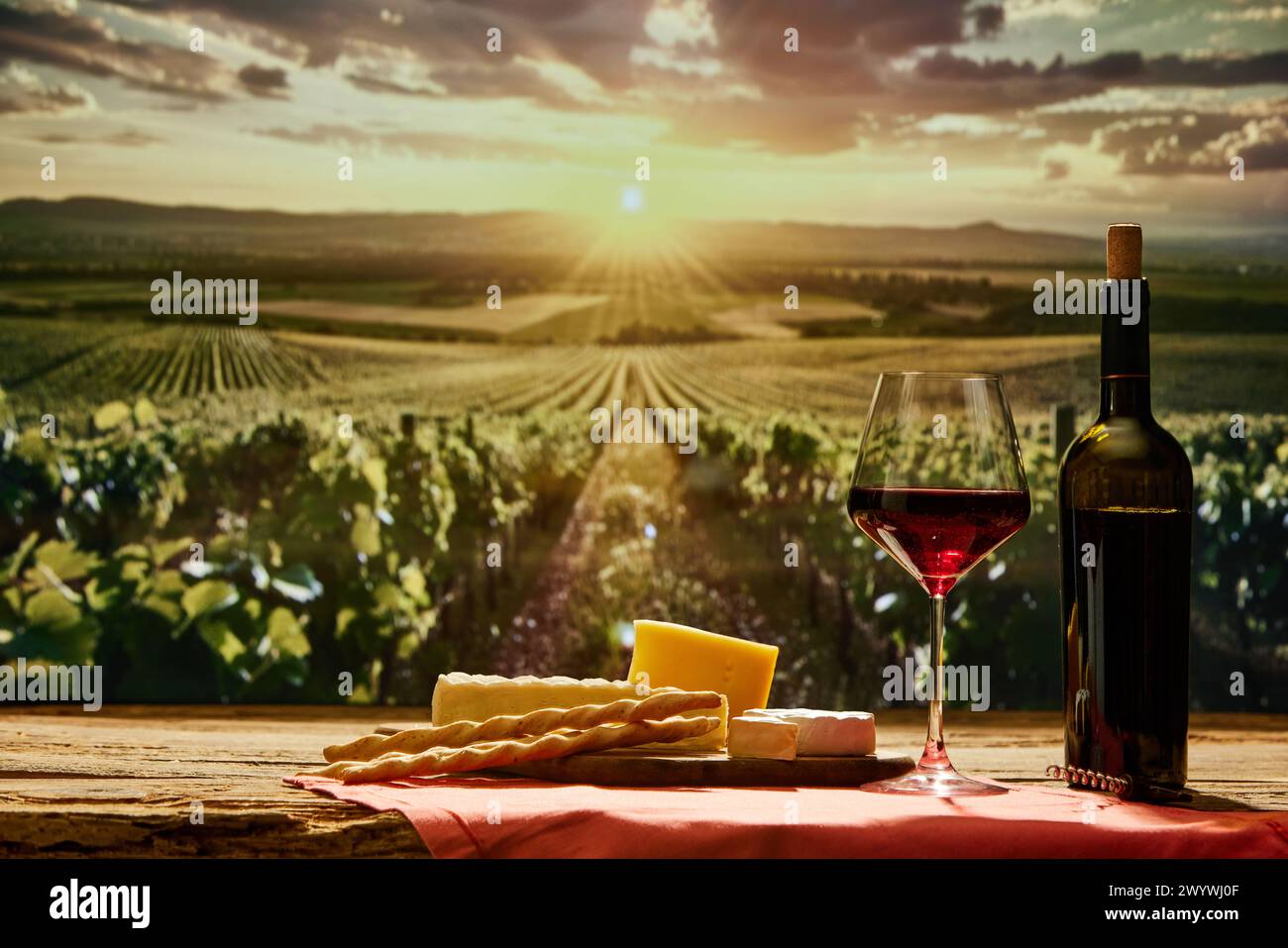 Glas Rotwein und Flasche auf Holztisch mit Käse und Brotstangen mit Weinberg während Sonnenuntergang im Hintergrund Stockfoto