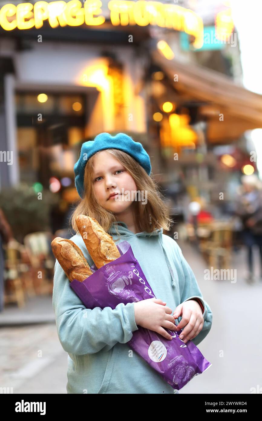 Porträt eines hübschen jungen französischen Teenagers in blauer Baskenmütze, der Baguettes vor dem Hintergrund der Abendlichter eines Cafés oder einer Bäckerei auf einem Narr hält Stockfoto