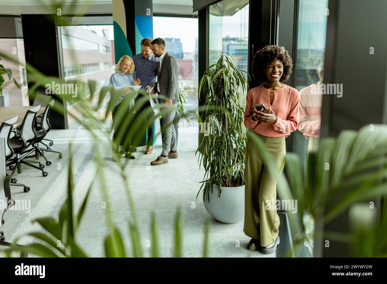 Die afroamerikanische Frau am Telefon steht neben üppigen Pflanzen im Haus, während sich die Kollegen im Hintergrund unterhalten Stockfoto