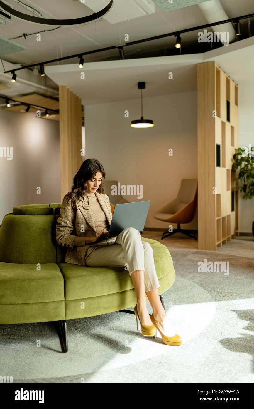 Eine fokussierte Frau arbeitet an ihrem Laptop, sitzt auf einem schicken grünen Sofa in einem gut beleuchteten modernen Büro Stockfoto