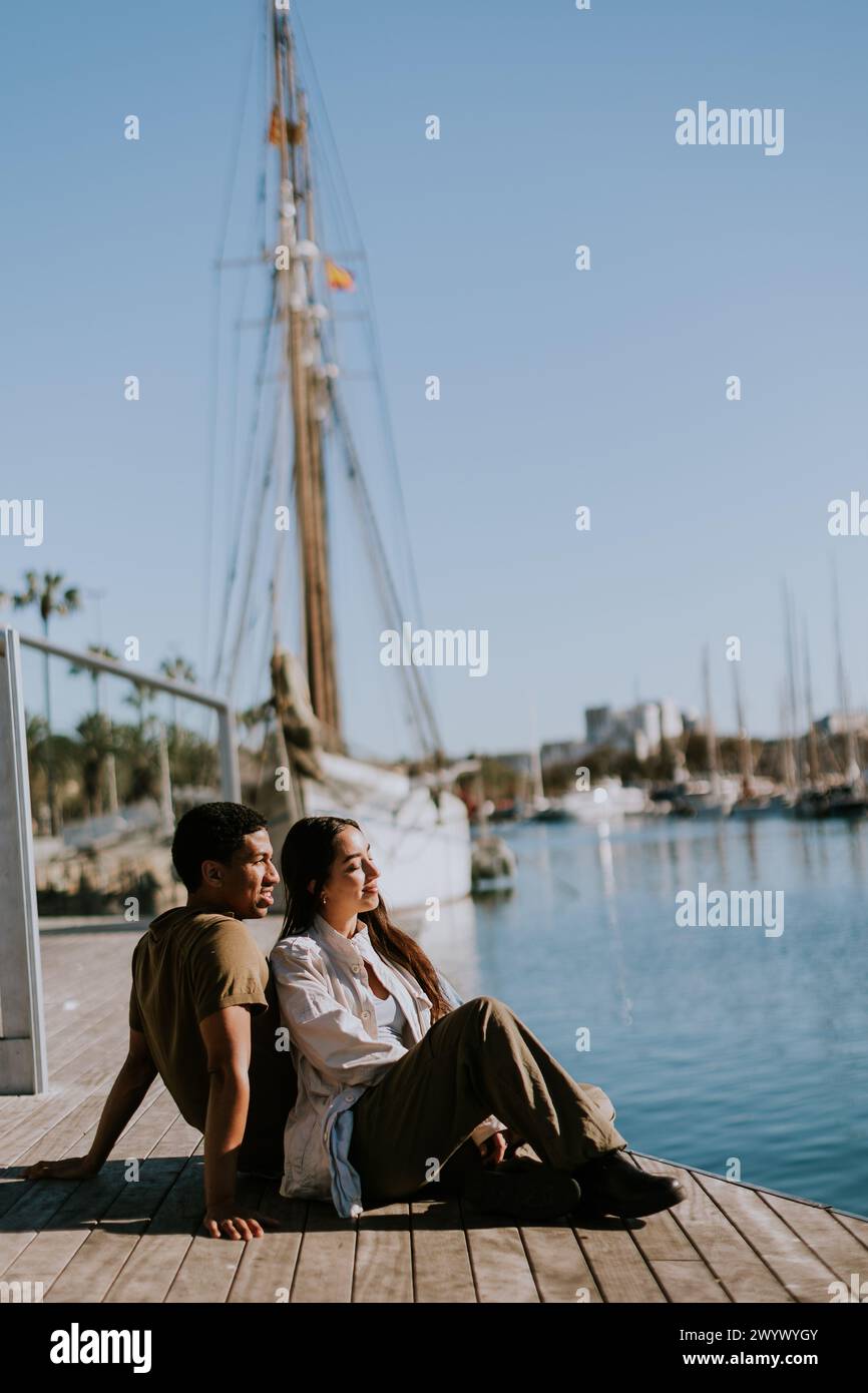 Ein Paar sitzt nahe an einem Dock und genießt einen ruhigen Moment mit Blick auf den Yachthafen an einem sonnigen Tag in Barcelona. Stockfoto