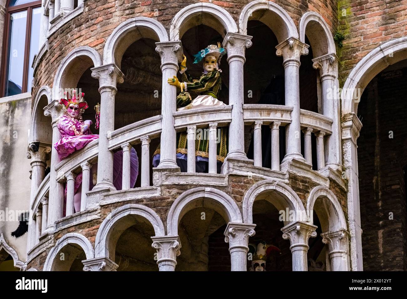 Zwei weibliche maskierte Personen in kreativen Kostümen posieren auf der Treppe der Scala Contarini del Bovolo, um den venezianischen Karneval zu feiern. Stockfoto